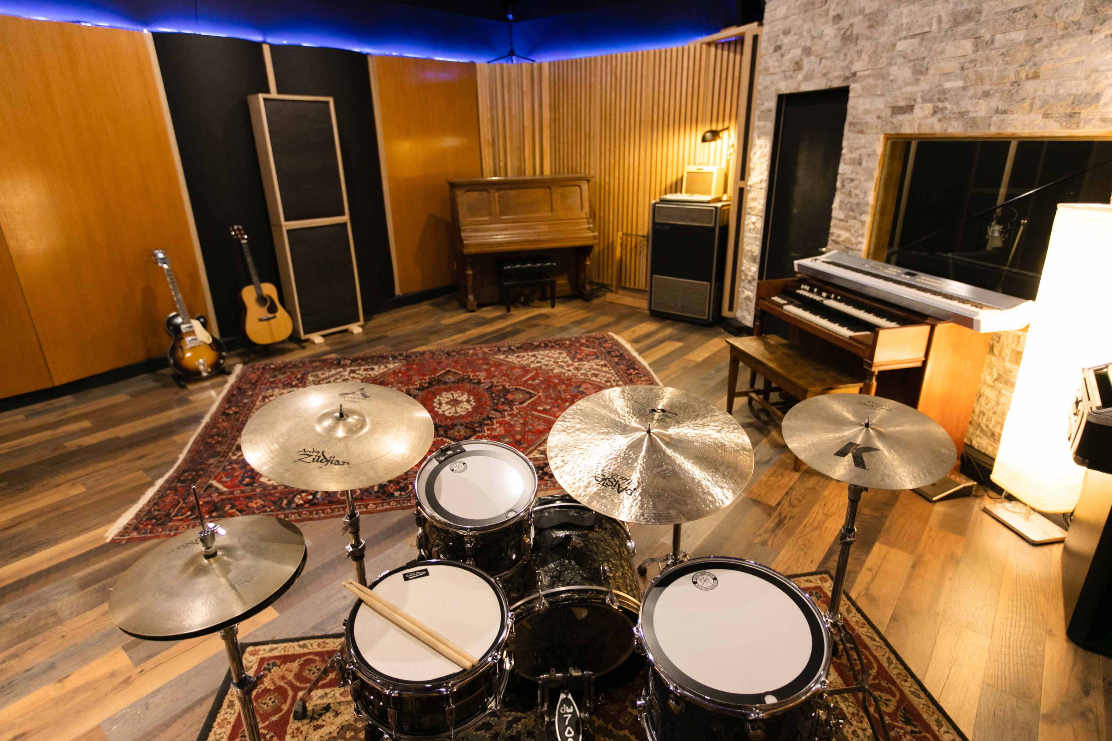 A music studio featuring a drum set in the foreground, a piano and a vintage amplifier in the background, and acoustic treatment on the walls.