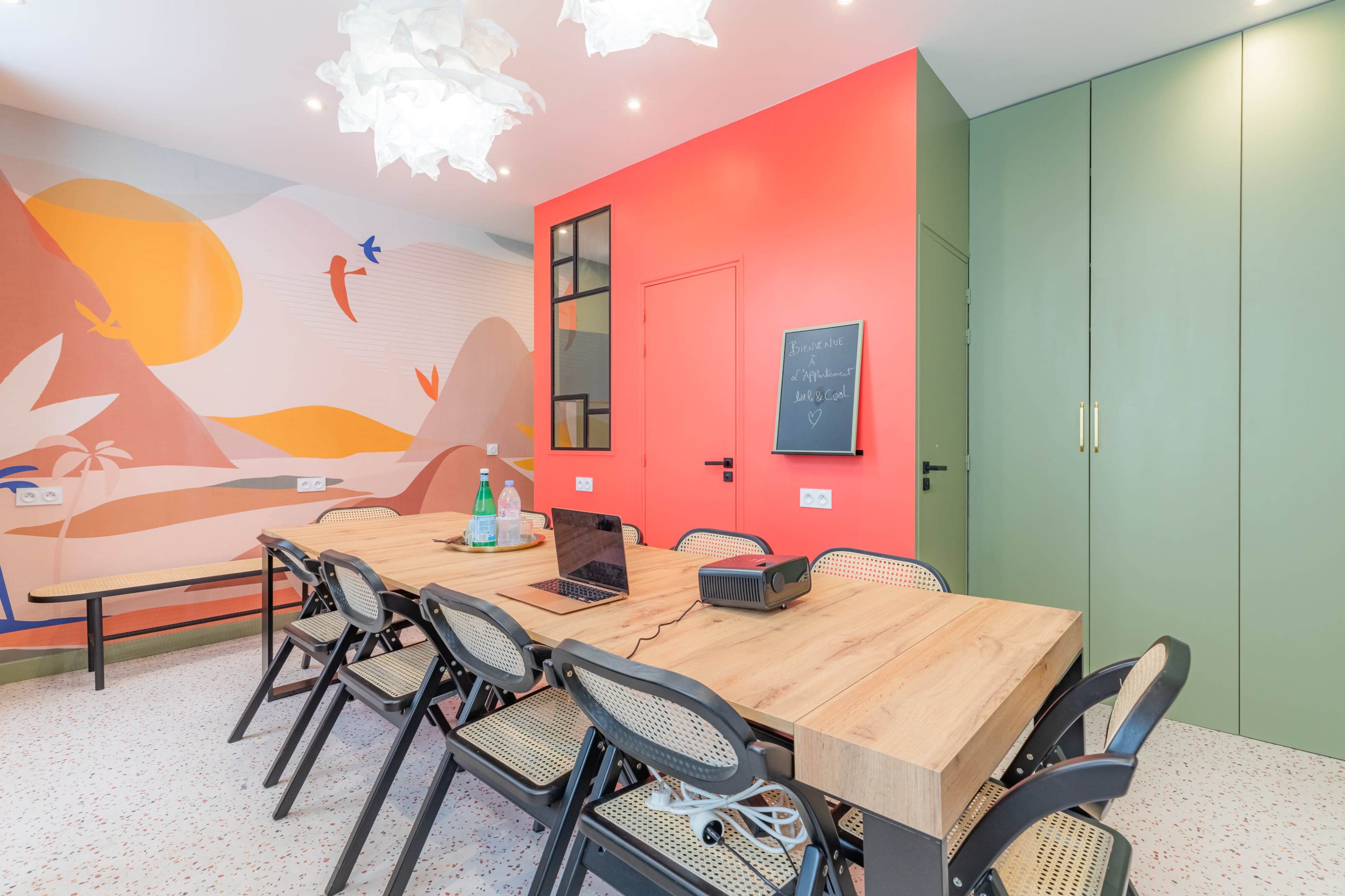A brightly colored meeting room features a long wooden table surrounded by black folding chairs, with a large mural on one wall and green cabinetry on another.