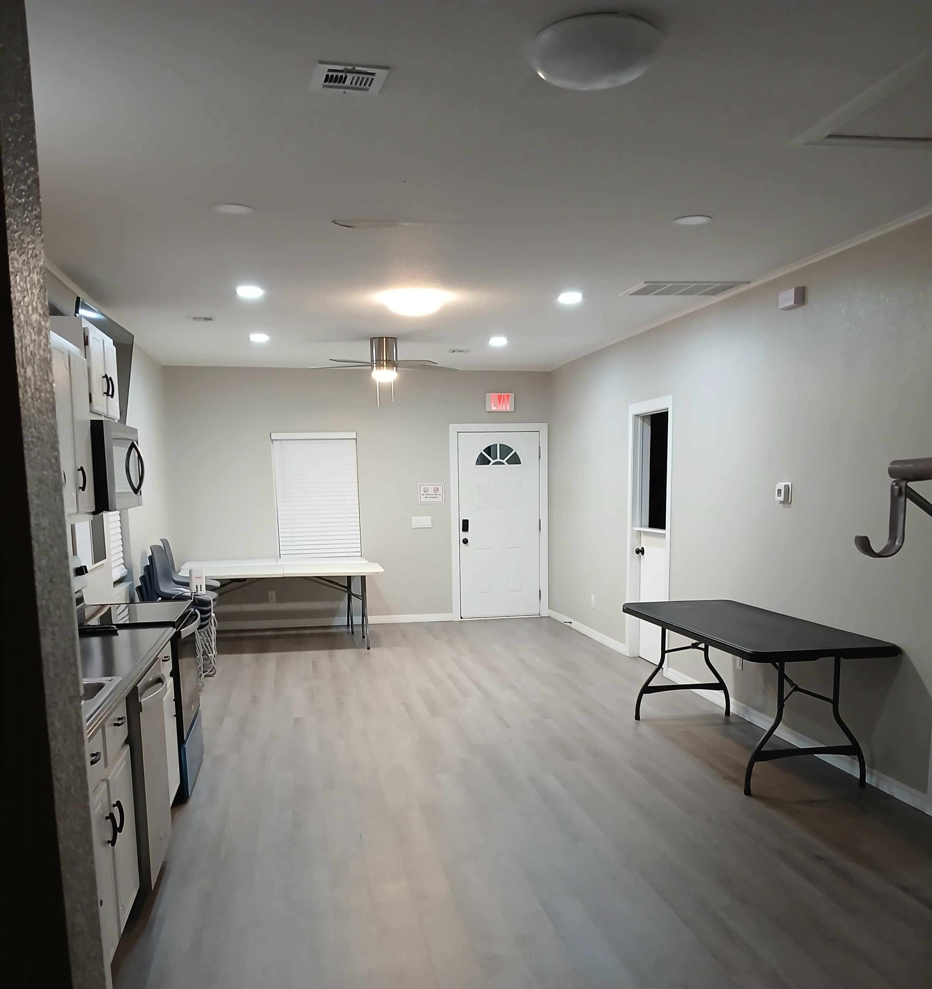 The image shows a spacious, well-lit room with a kitchen area on one side, a table in the center, and a front door opposite the kitchen.