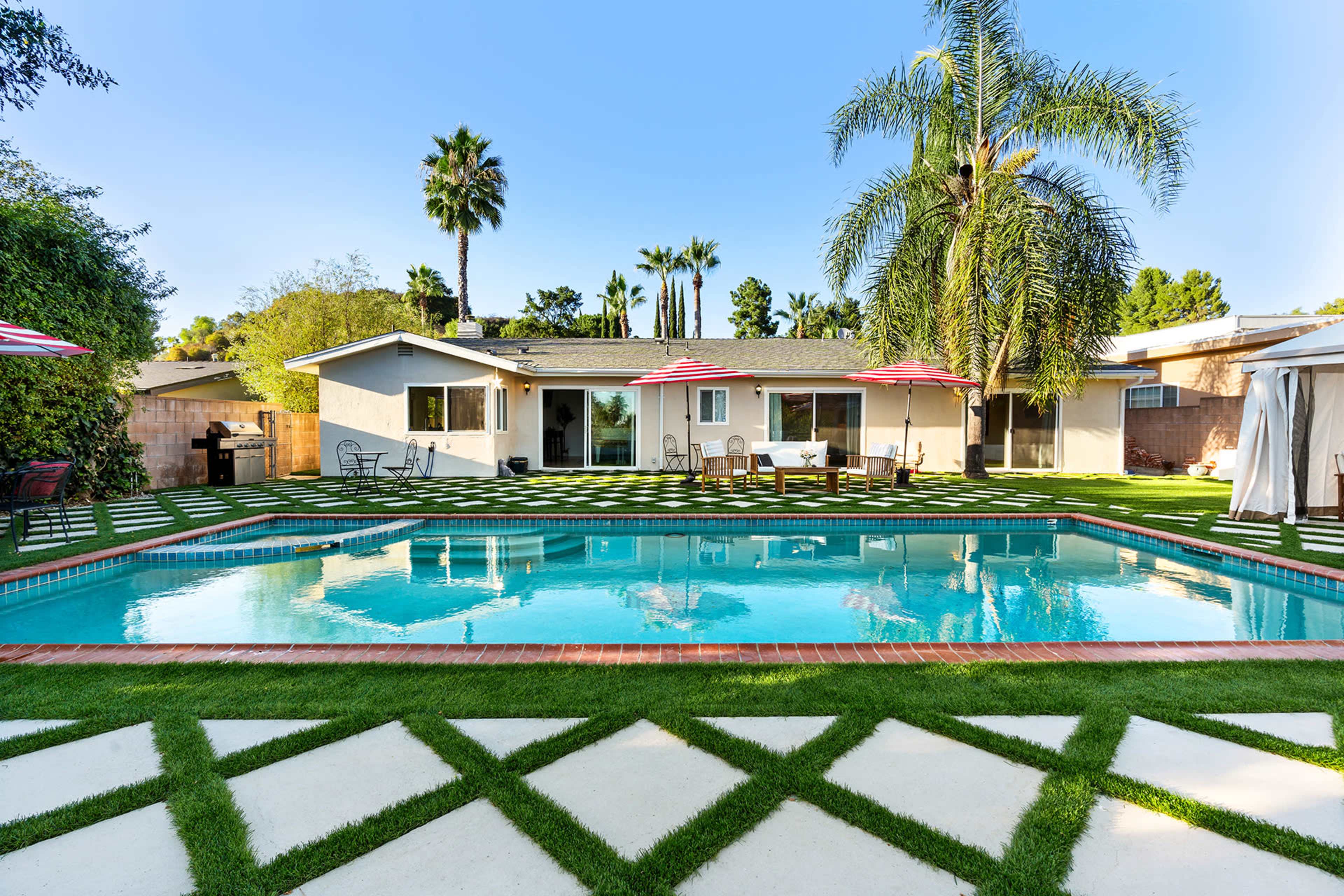 The image shows a backyard with a swimming pool surrounded by green grass and a tiled patio, alongside a single-story house and palm trees.