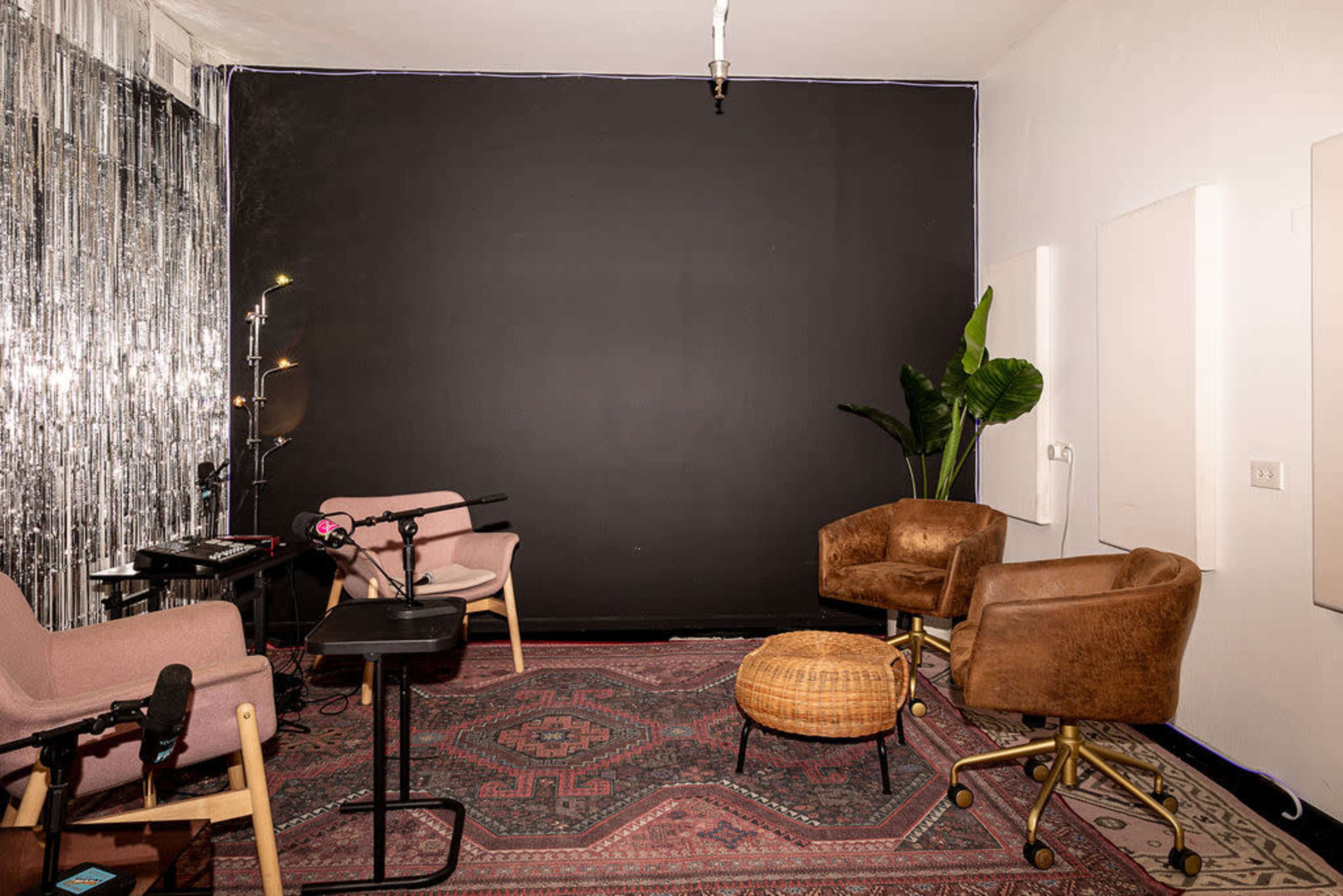 The image shows a small recording studio or podcast space featuring two pink chairs, a woven pouf, a black backdrop, and decorative elements like a plant and a metallic curtain.