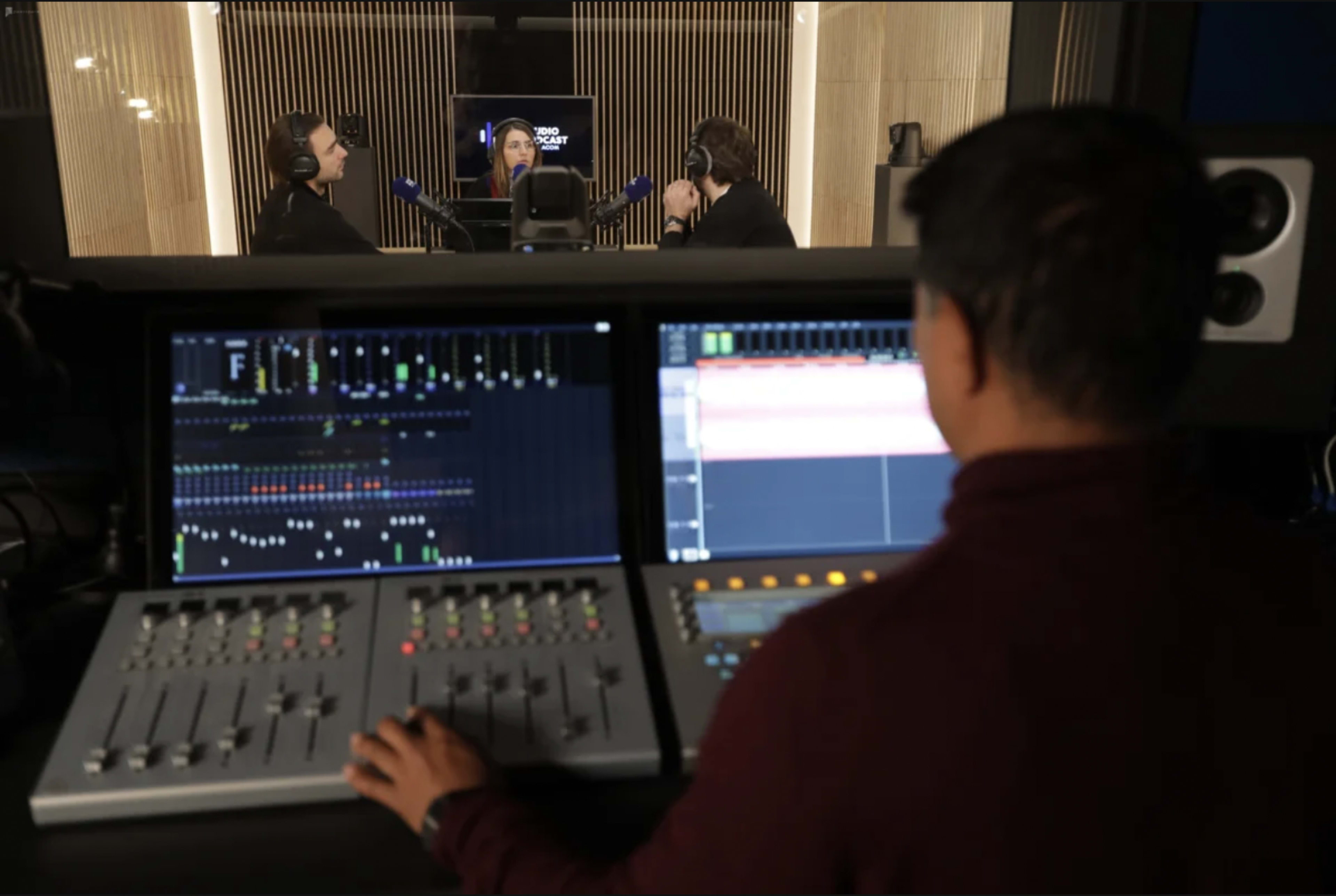 A sound engineer monitors audio levels while two individuals engage in conversation in a recording studio.