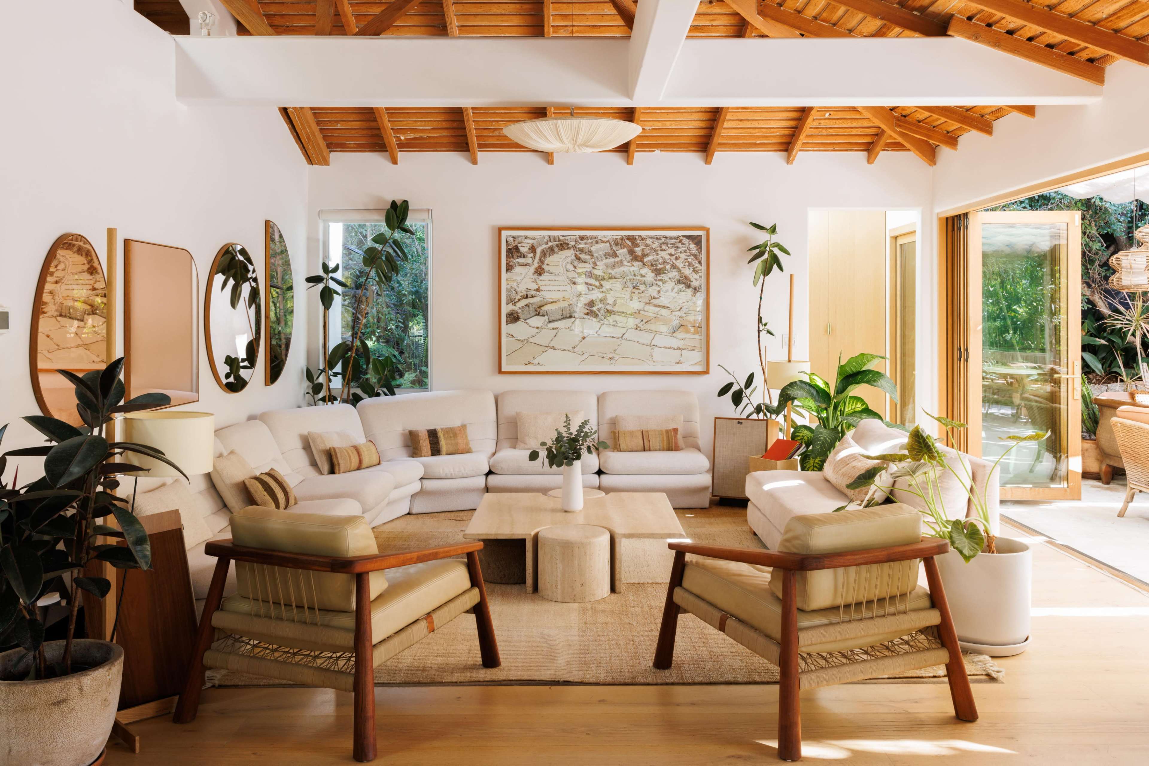 The image shows a modern living room with a high wooden ceiling, featuring white sofas, a light-colored coffee table, potted plants, and large windows opening to an outdoor area.