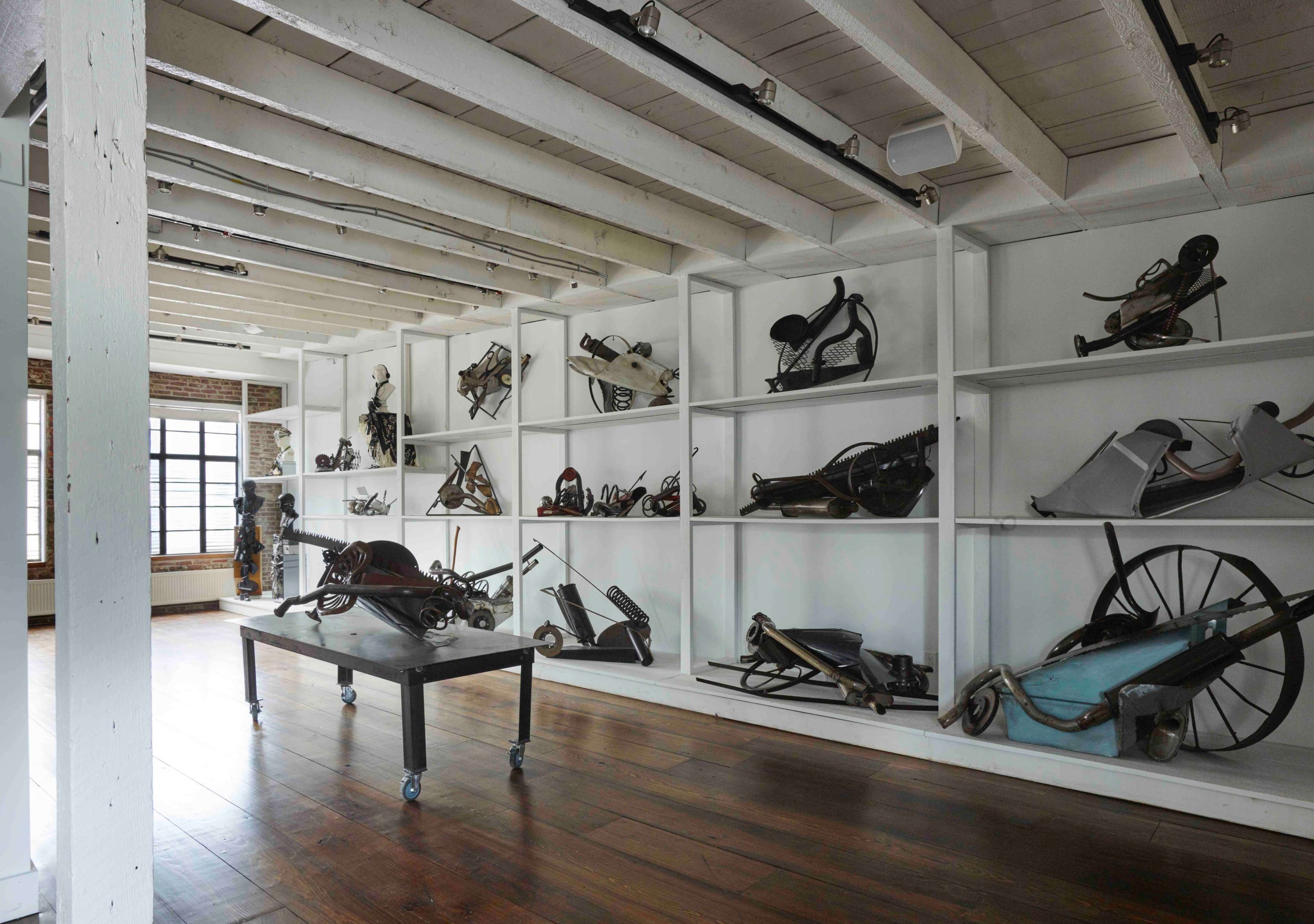The image shows a spacious room with wooden floors and white shelves displaying various antique industrial tools and equipment.