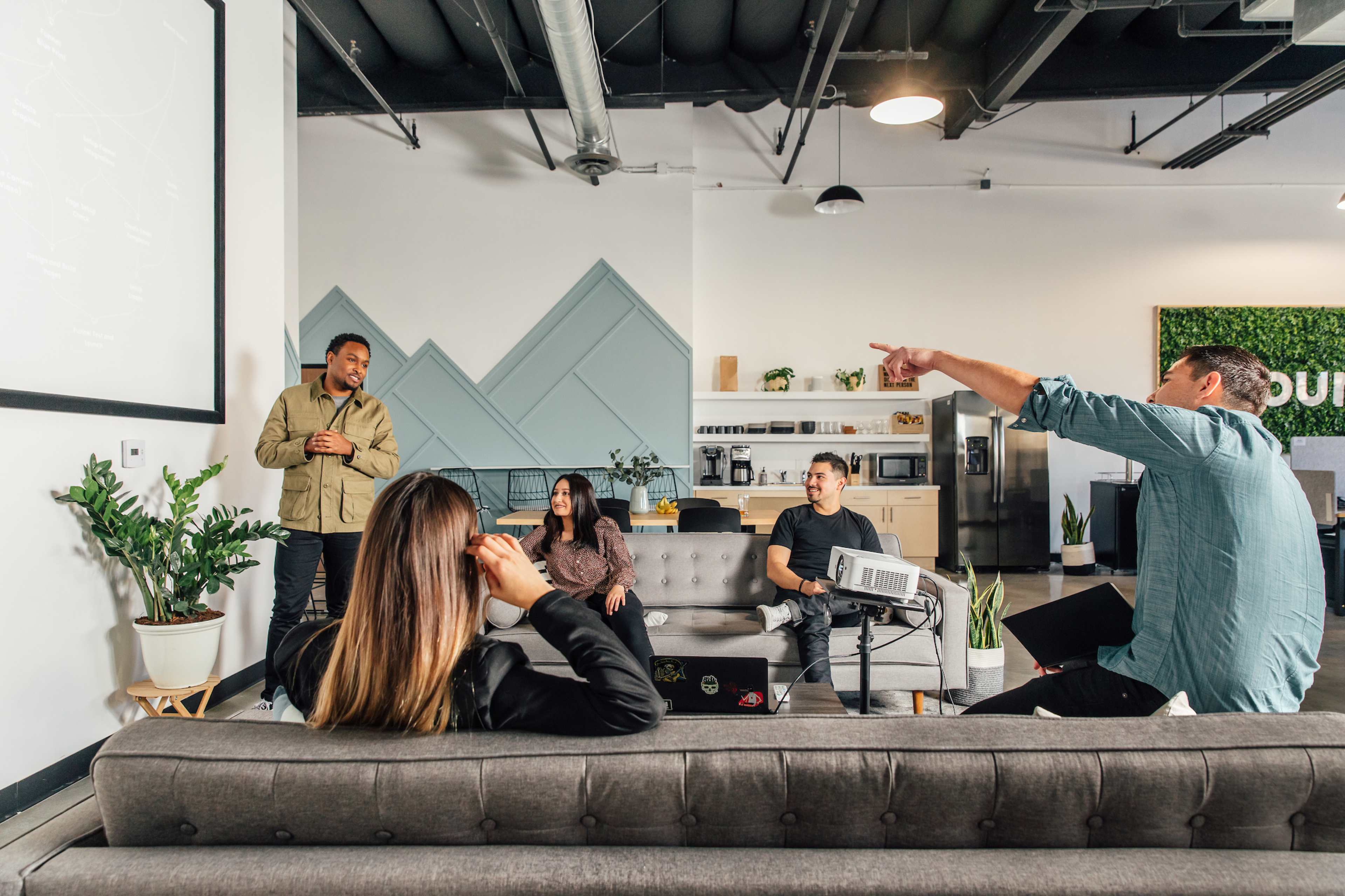 A group of four people are engaged in a discussion in a modern office space, with one person standing and presenting while the others listen from a couch.