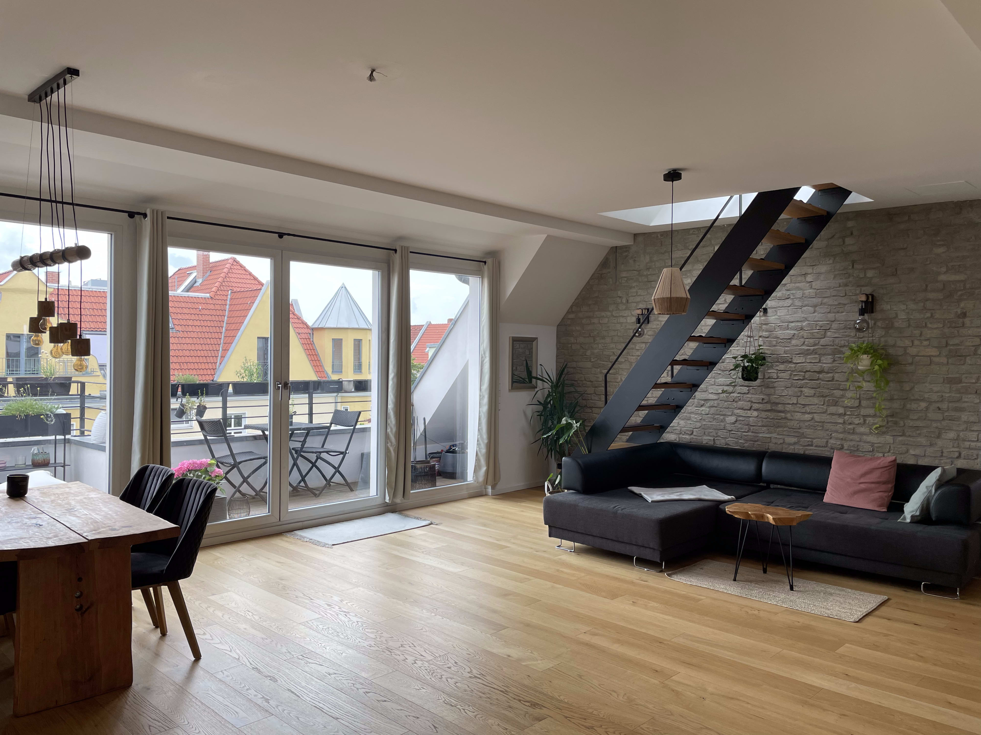 A modern living room with a wooden dining table, a sectional sofa, and large windows overlooking a balcony with a view of rooftops.