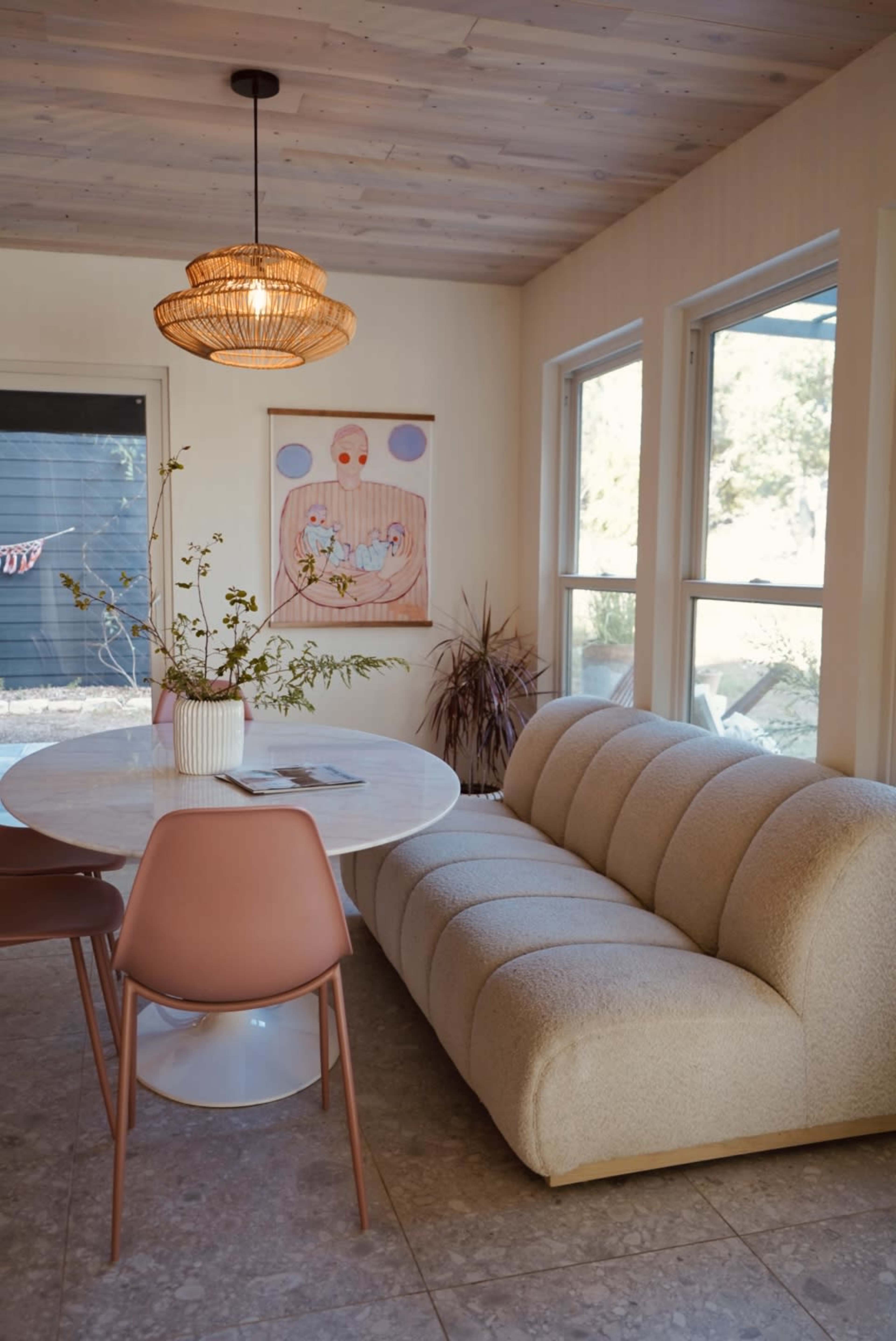 A cozy dining area features a round white table, a textured beige sofa, and large windows that let in natural light.