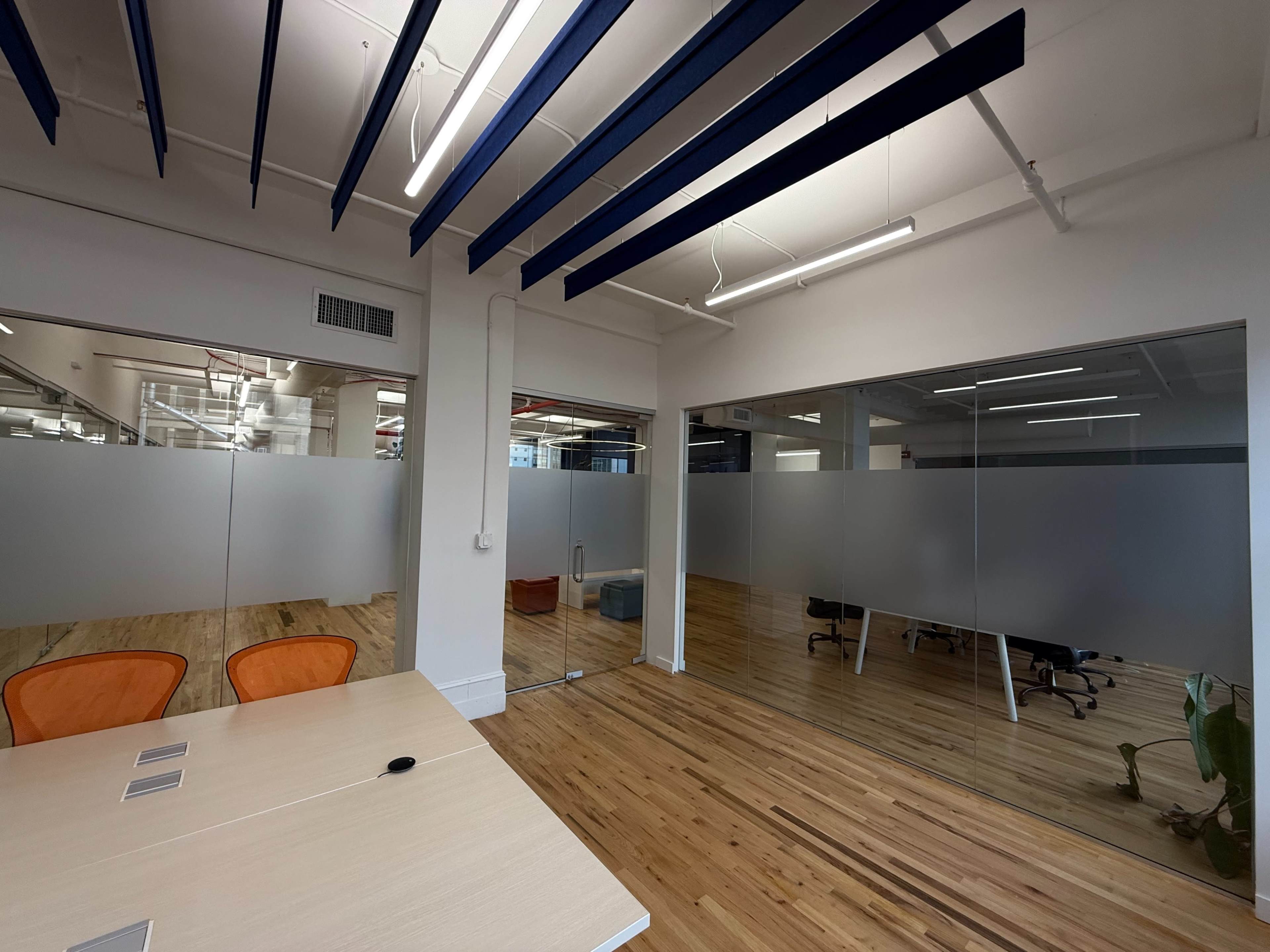 The image shows a modern office space with wooden flooring, glass-walled meeting rooms, and a conference table with orange chairs.