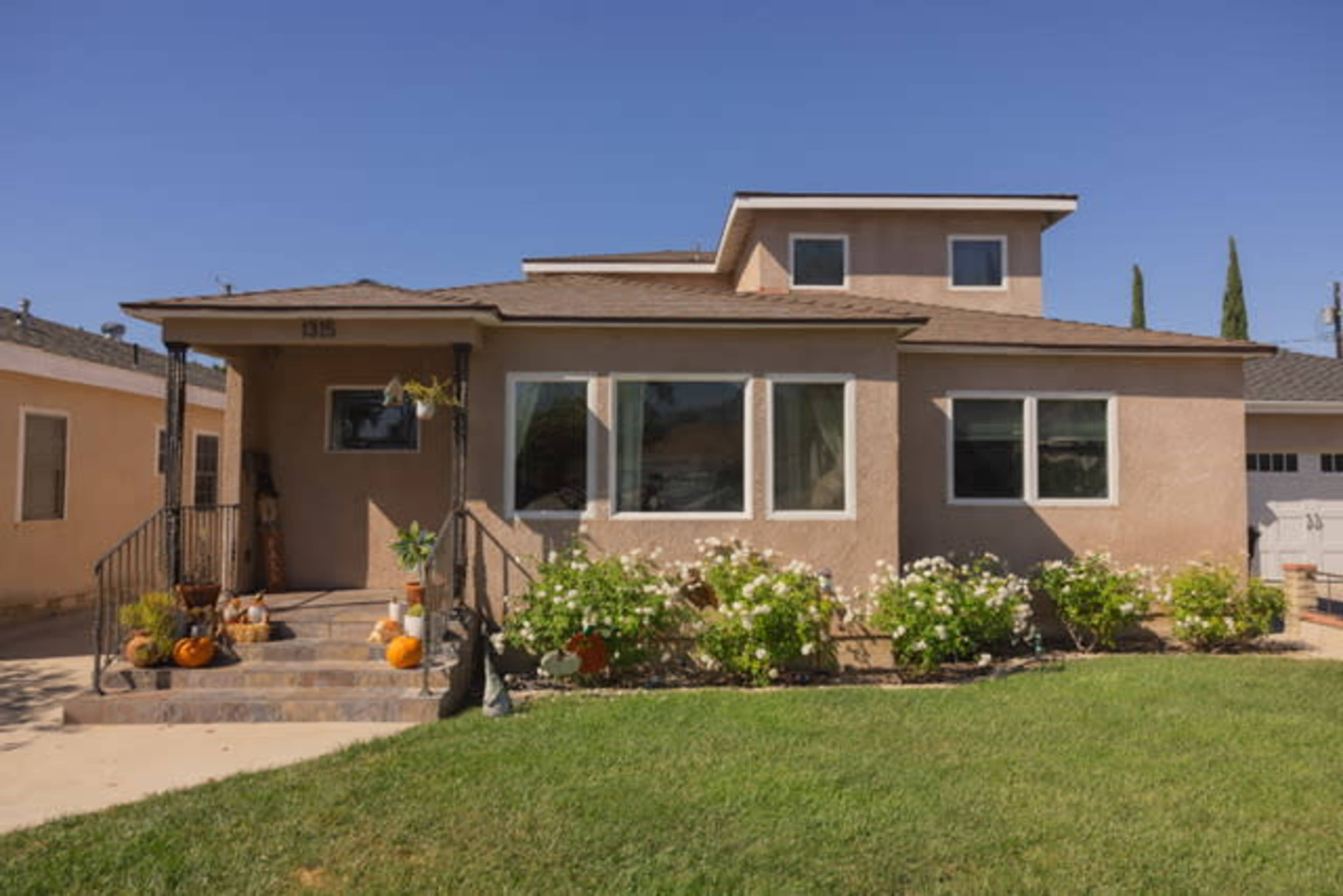 A single-story house with a flat-roofed upper section, a front porch, and a well-maintained garden featuring several pumpkins and shrubs.