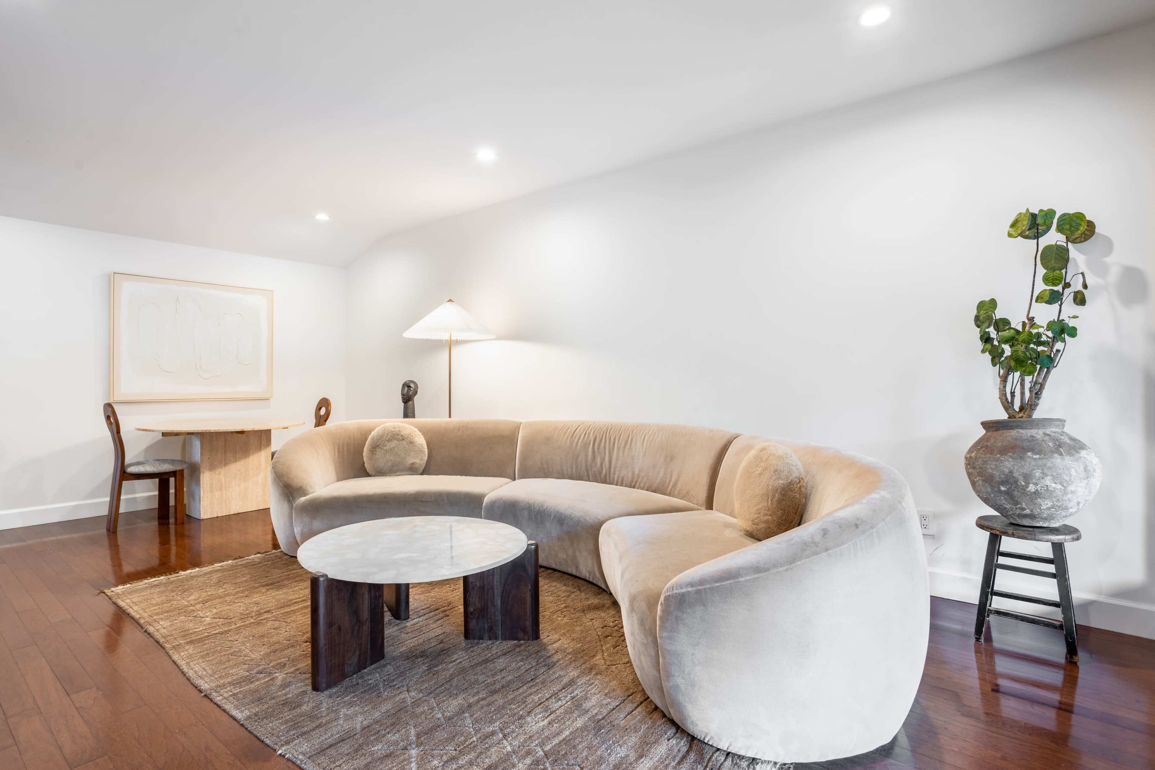 A modern living room featuring a curved beige sofa, a circular marble-top coffee table, a wooden desk, a lamp, and a potted plant in a decorative pot.