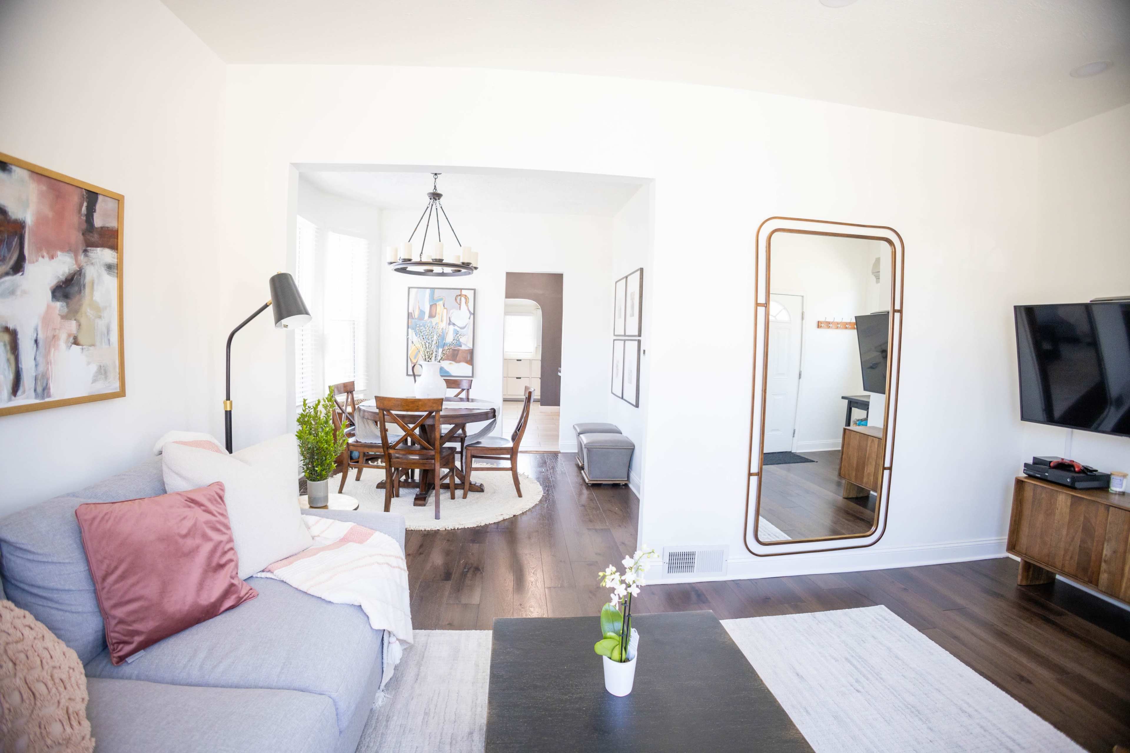 A living room features a gray couch with pillows, a black coffee table, and a dining area visible in the background with a round table and chairs.