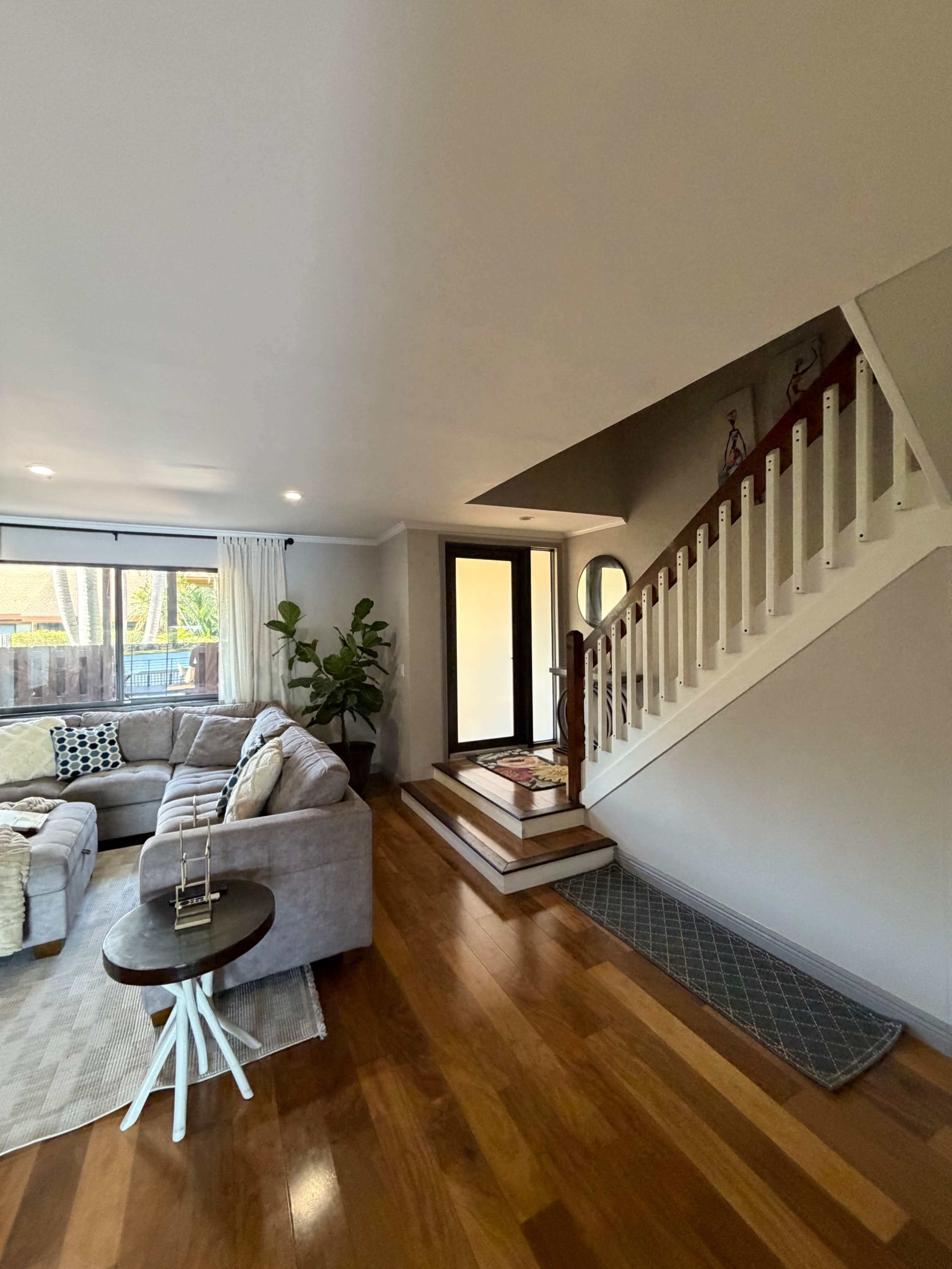 The image shows a cozy living room with a gray sectional sofa, a staircase leading to an upper level, and a large entrance door at the back.