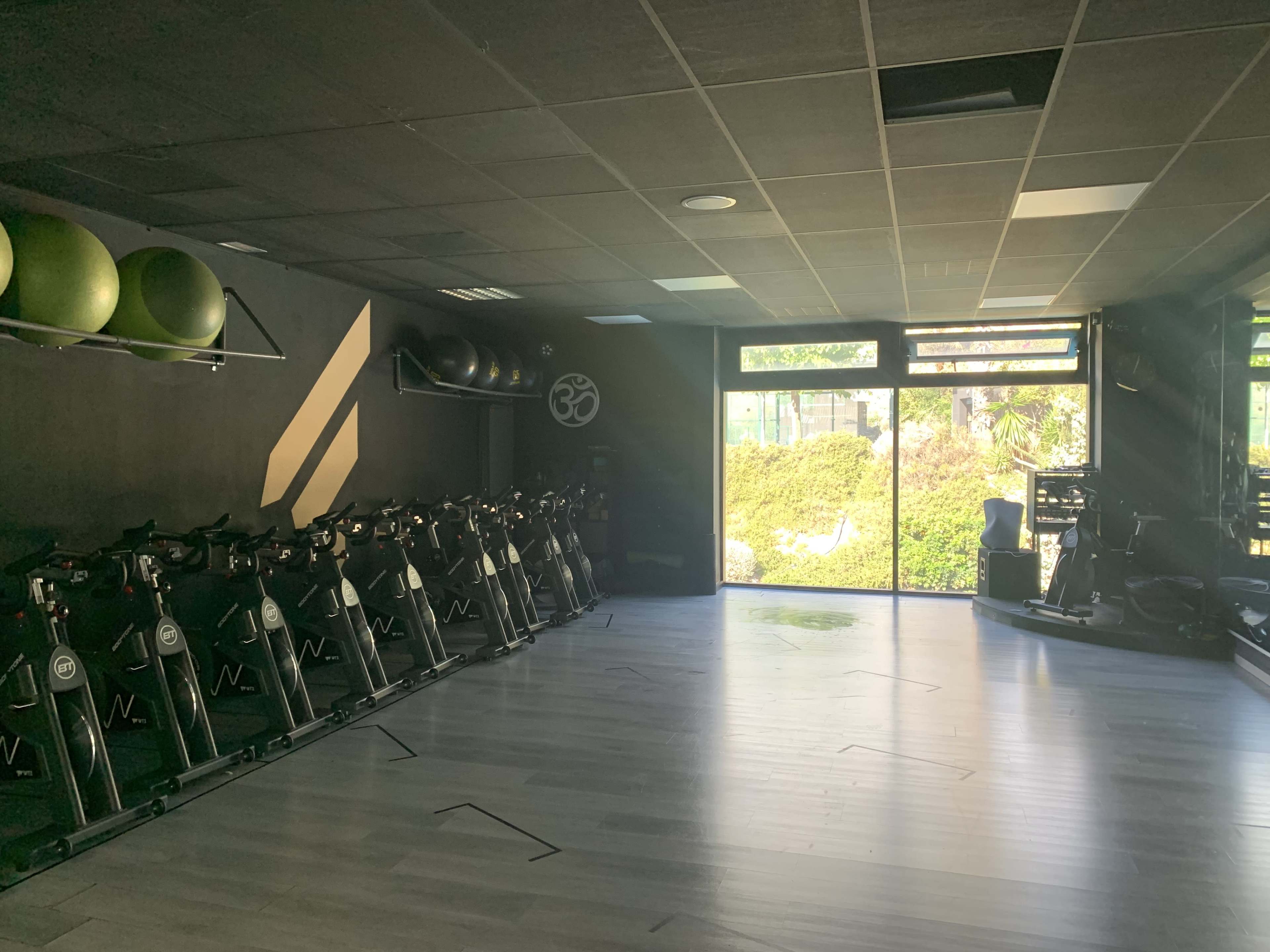 A fitness studio with exercise bikes lined up along one wall and fitness balls stored on another wall, illuminated by natural light coming through large windows.