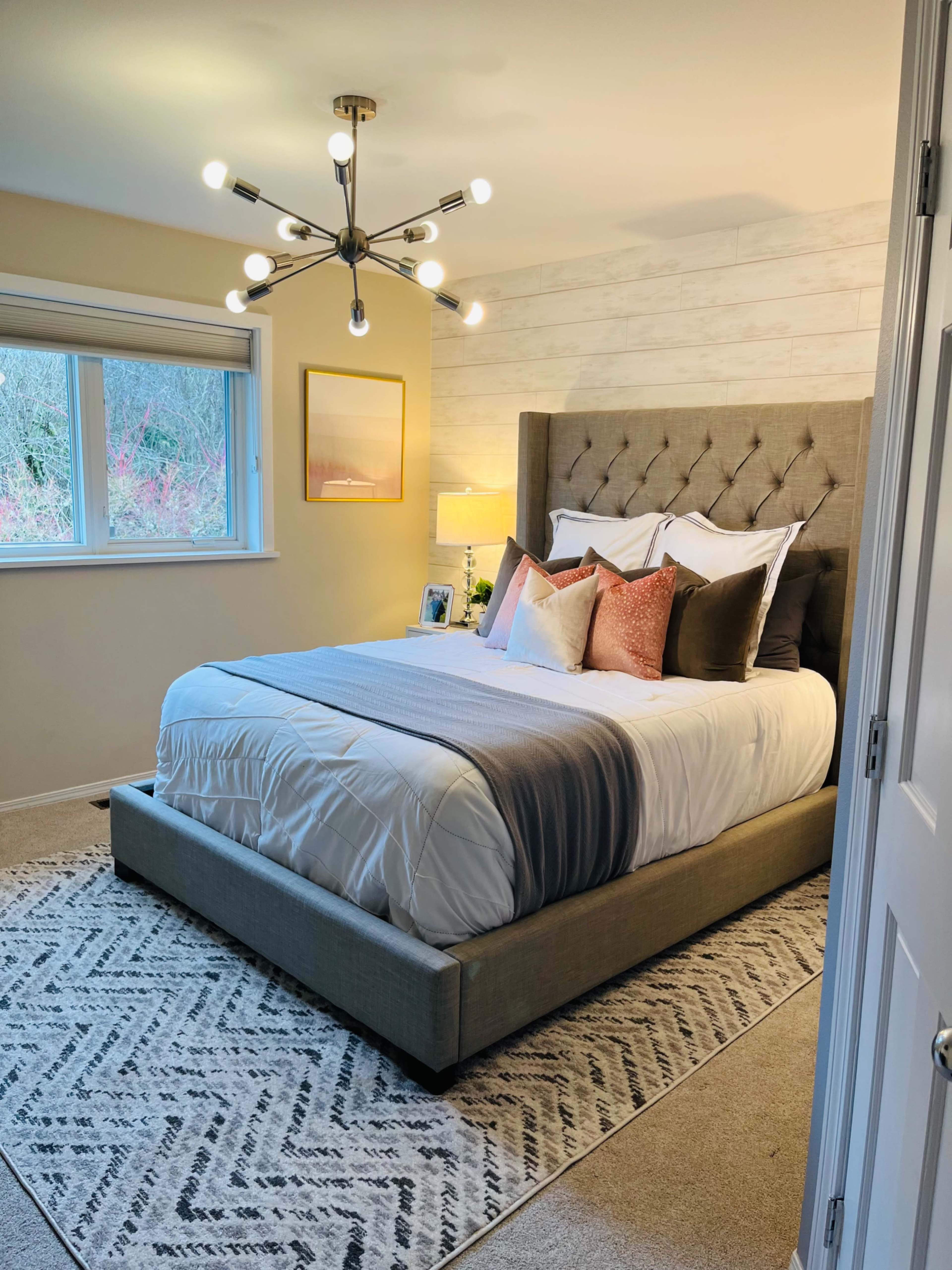 A well-lit bedroom featuring a tufted bed with decorative pillows, a bedside lamp, and a modern light fixture hanging from the ceiling.