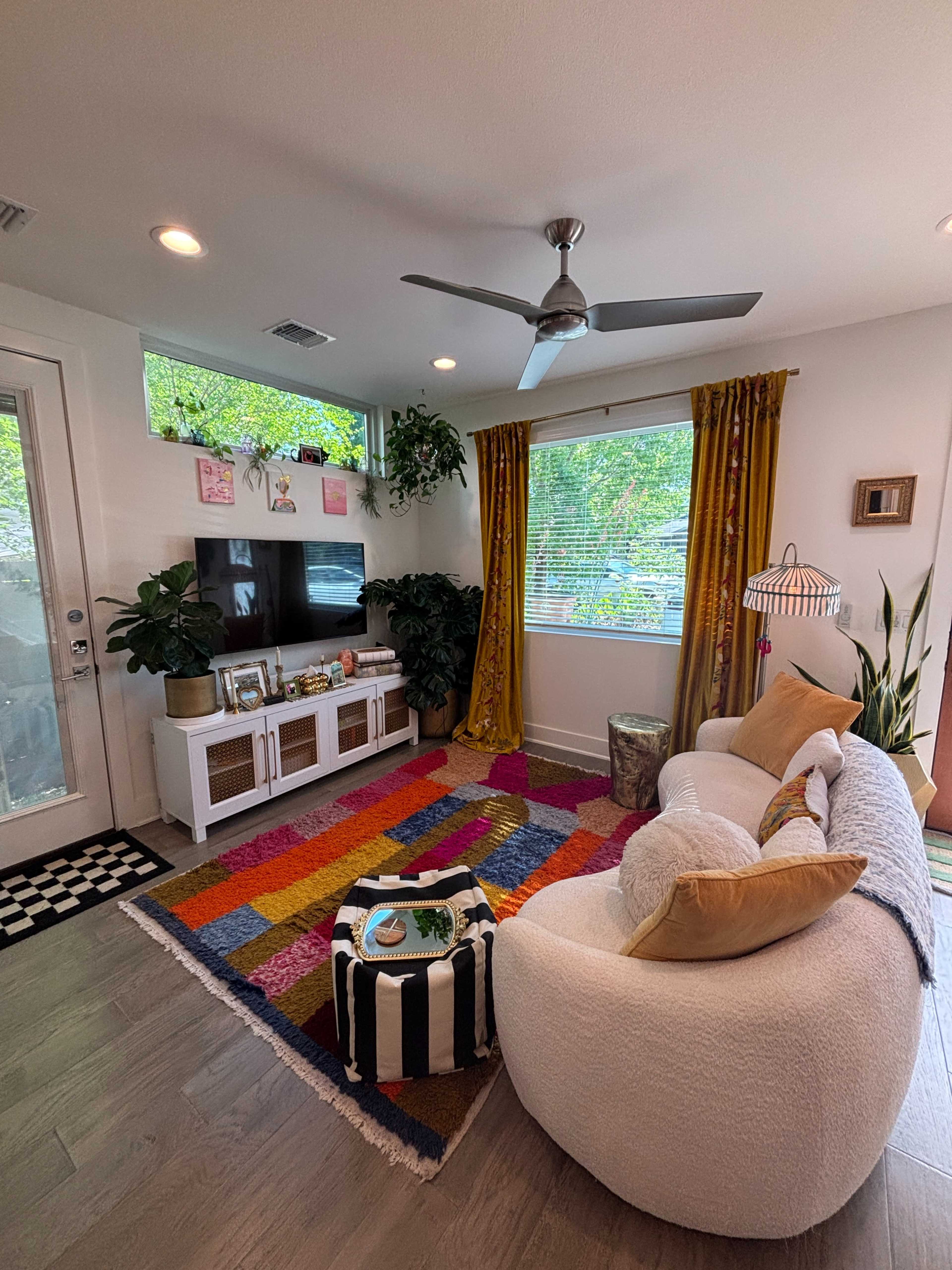 The image shows a modern living room featuring a large colorful rug, a white sofa with throw pillows, a TV stand, and windows adorned with yellow curtains, alongside plants and decor elements.