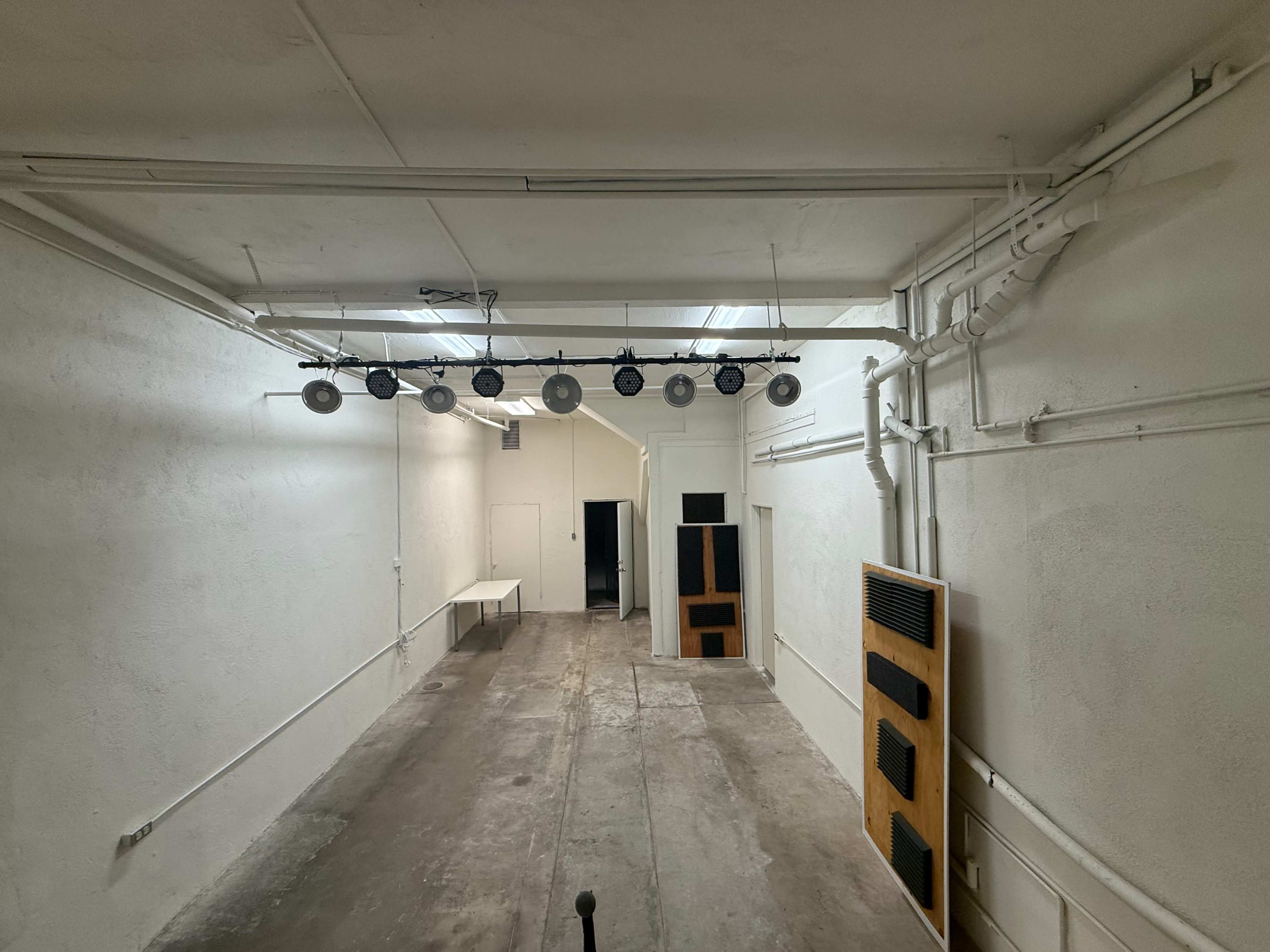 The image shows a spacious, empty room with white walls, overhead lights, and a concrete floor, featuring a table on one side and speakers mounted on the wall.