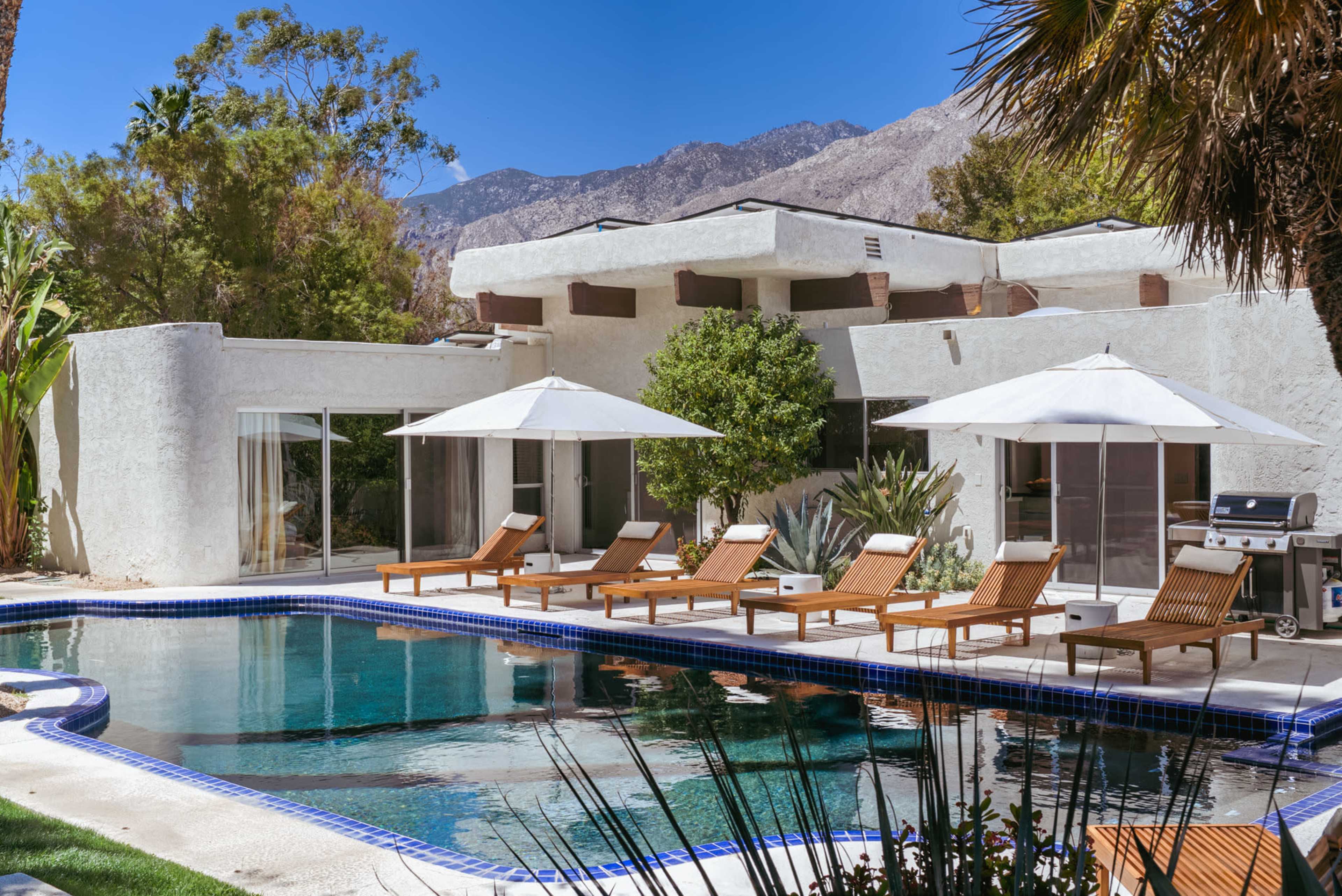 The image depicts a modern house with a swimming pool surrounded by lounge chairs and umbrellas, set against a backdrop of mountains and clear blue skies.