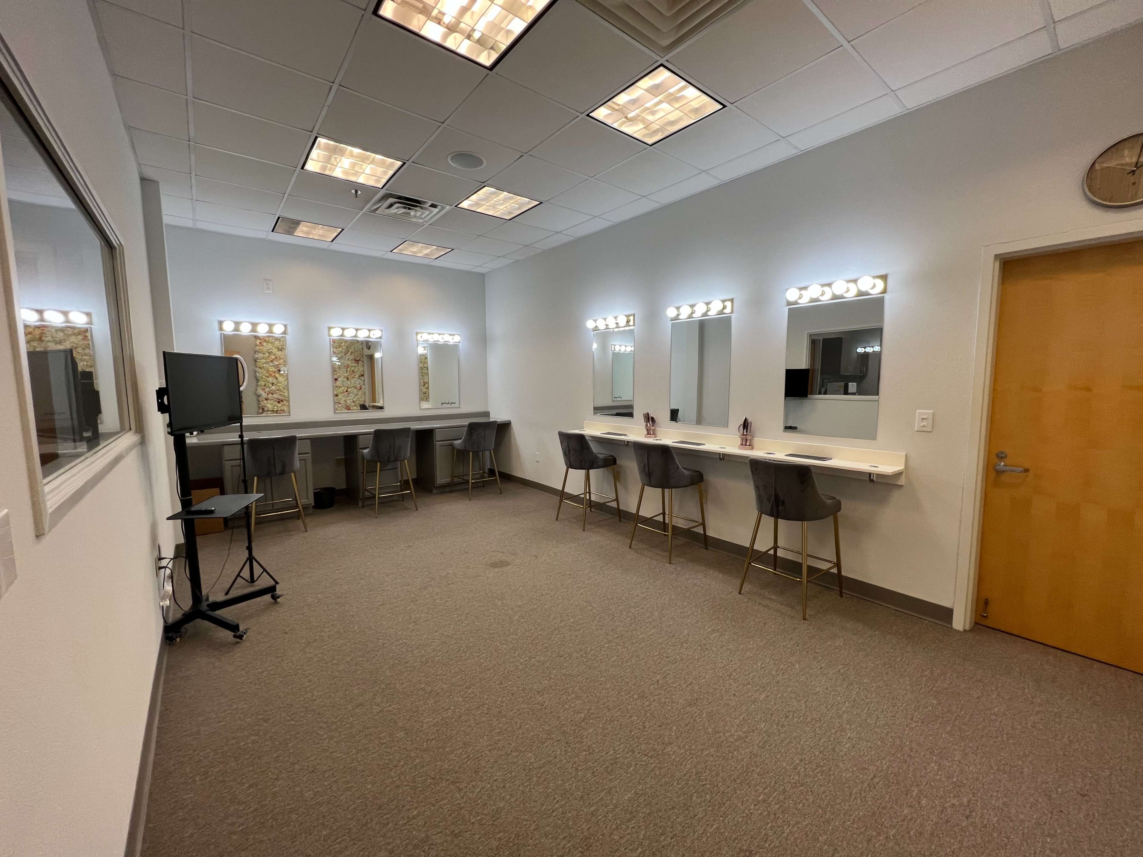 A brightly lit dressing room features five mirrors with lighted frames, a long white counter, and several chairs against a neutral wall.