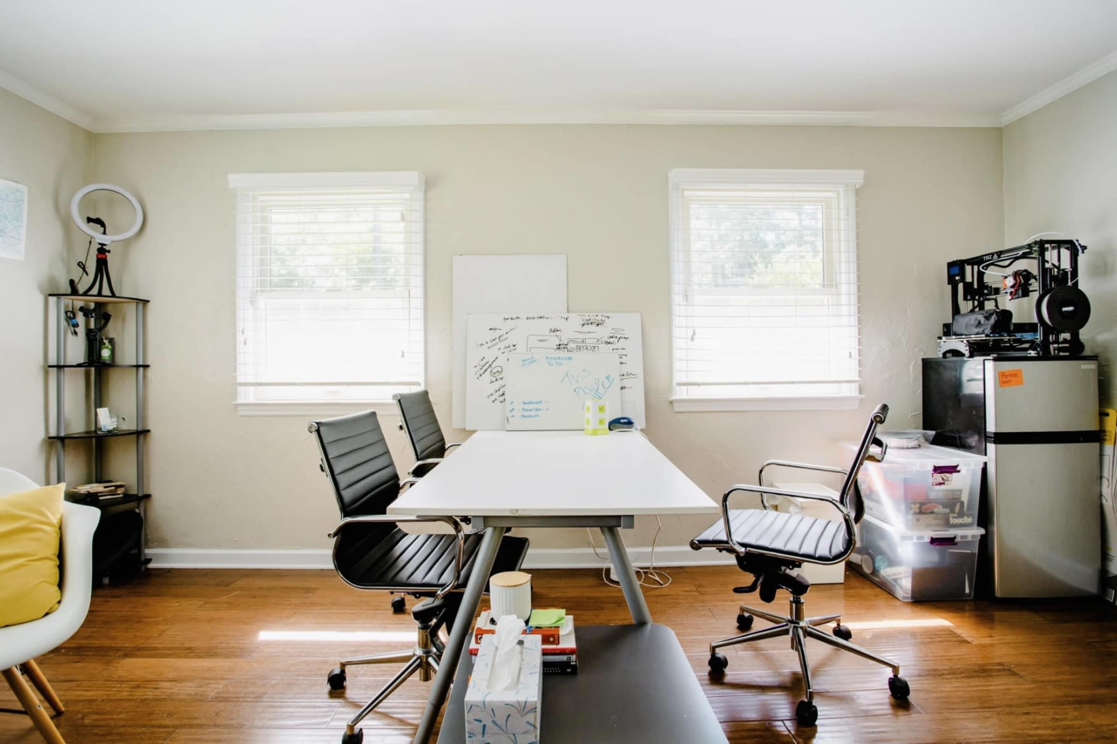 A bright, organized office space features a large table surrounded by black chairs, whiteboards, and storage units against the walls.