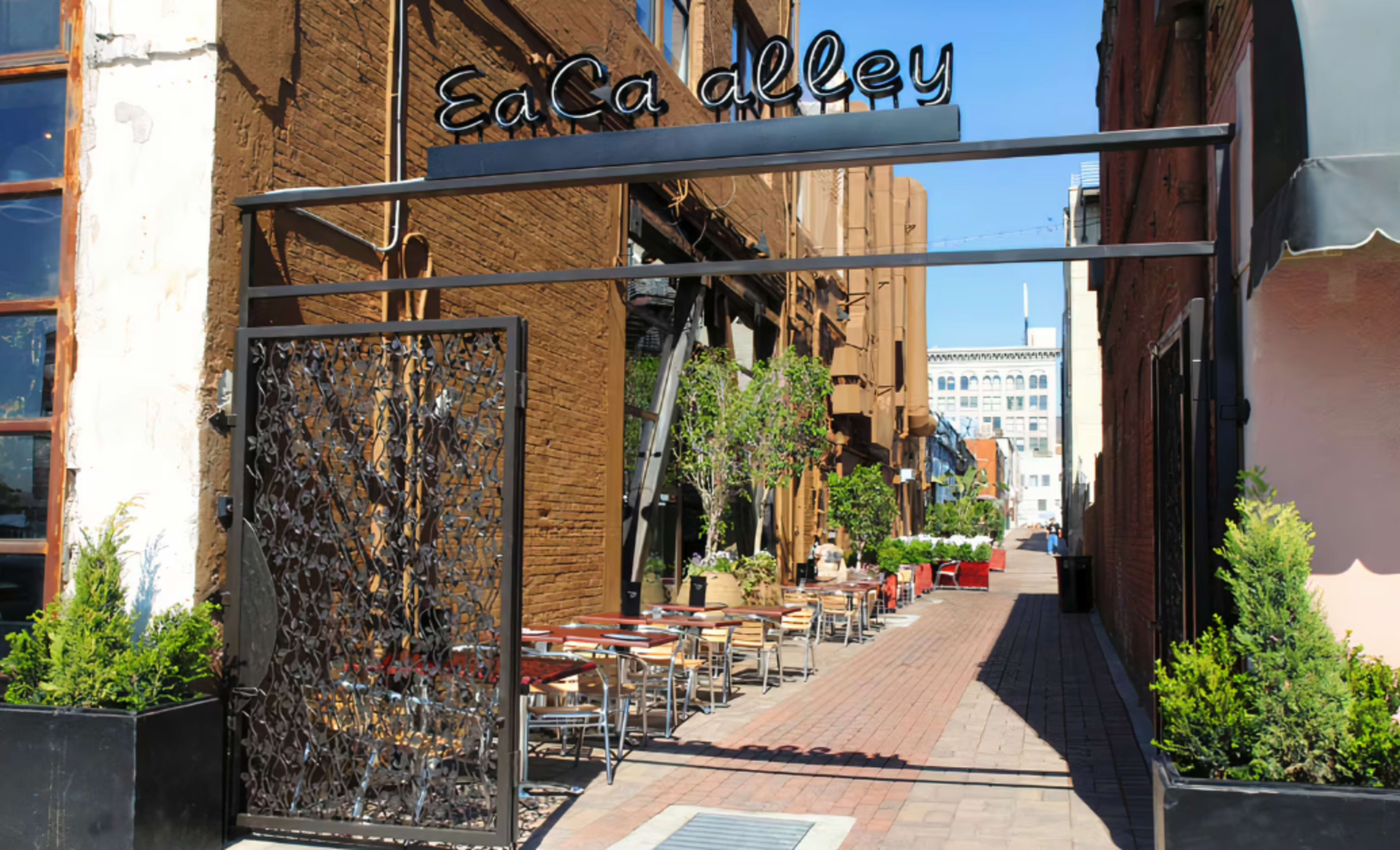 A narrow alleyway with outdoor seating is framed by brick buildings and features a decorative metal entrance sign that reads "EaCa Alley."