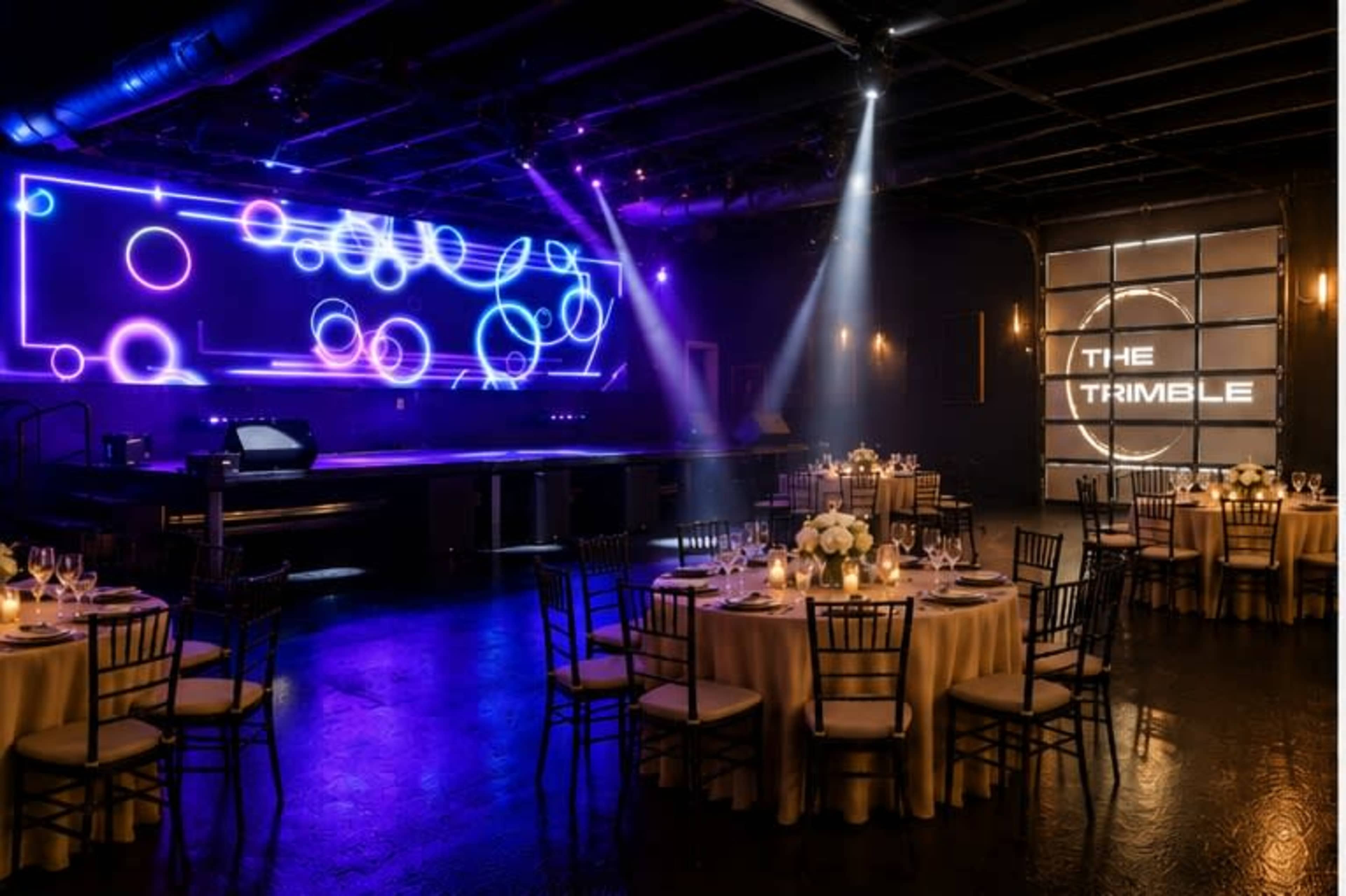 The image shows a dimly lit event space with round tables set for dining, illuminated by colorful light projections on a large screen and a wall that reads "THE TRIMBLE."