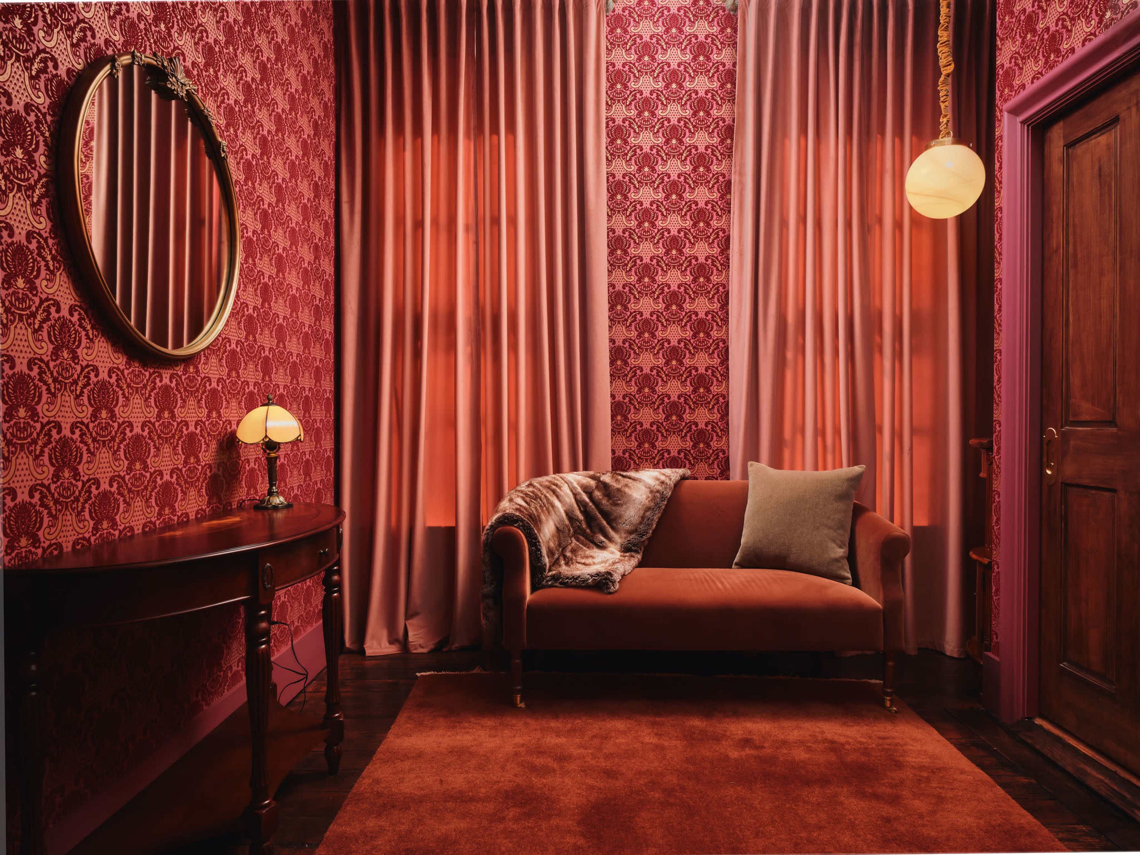 A velvet sofa with a decorative throw blanket is positioned against a wall adorned with patterned wallpaper, complemented by two elegant curtains and a round mirror.