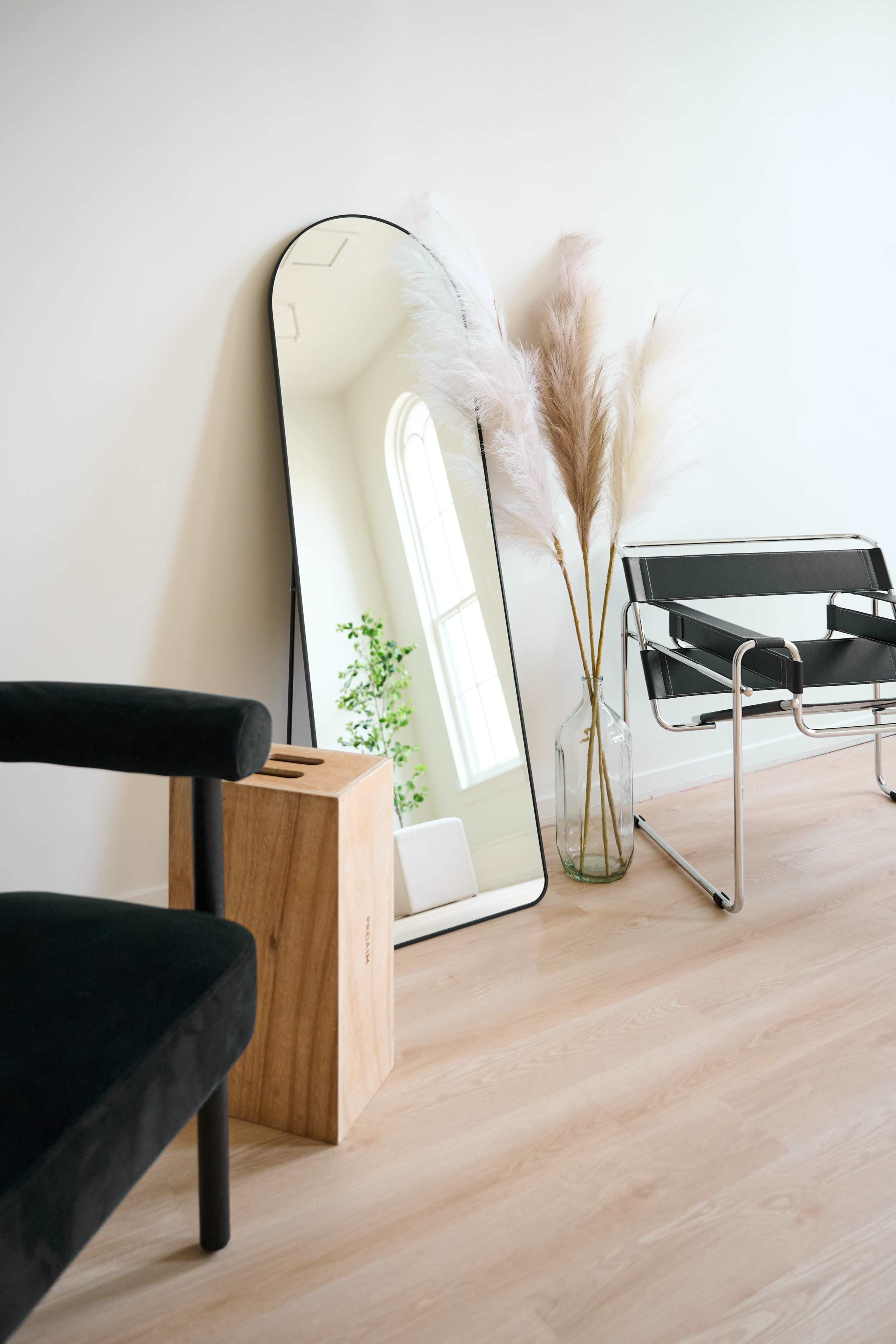 A tall mirror leans against a white wall beside a wooden stool and a glass vase with pampas grass, accompanied by a modern black chair.
