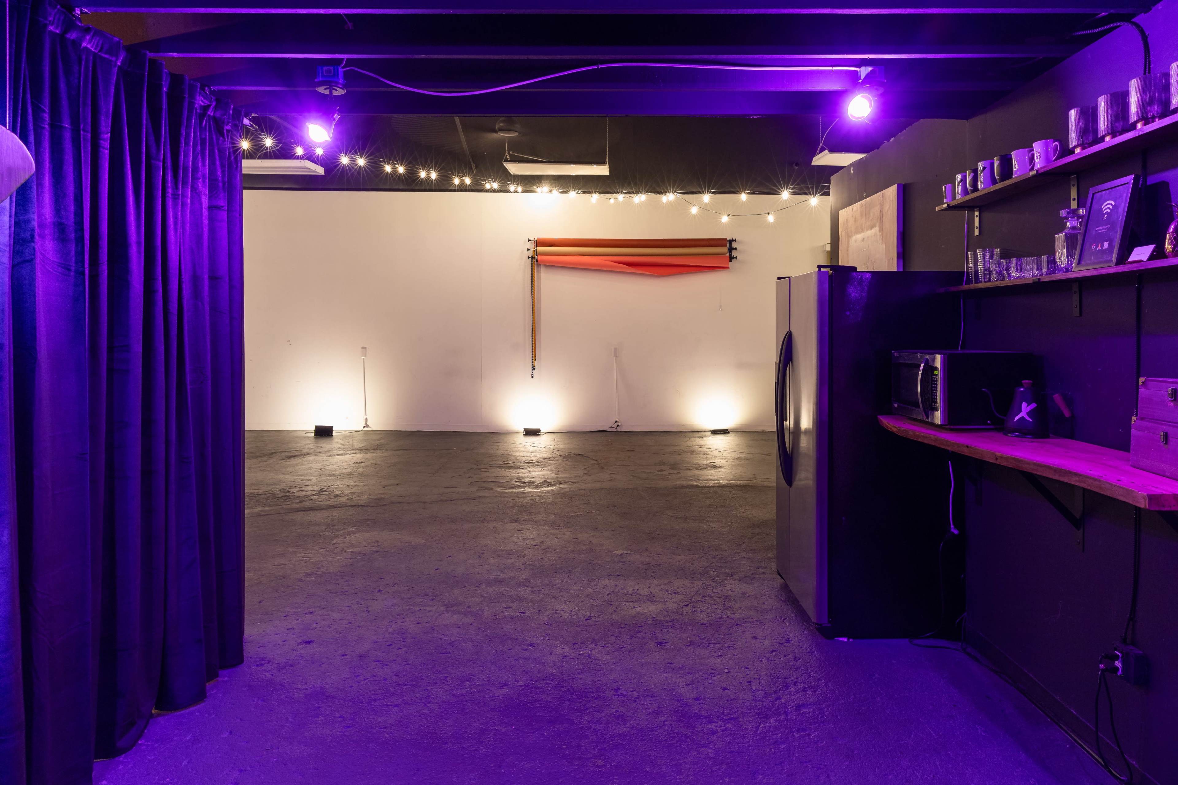The image shows a dimly lit, minimalist room with purple lighting, featuring a bare concrete floor and a wall adorned with a few objects.