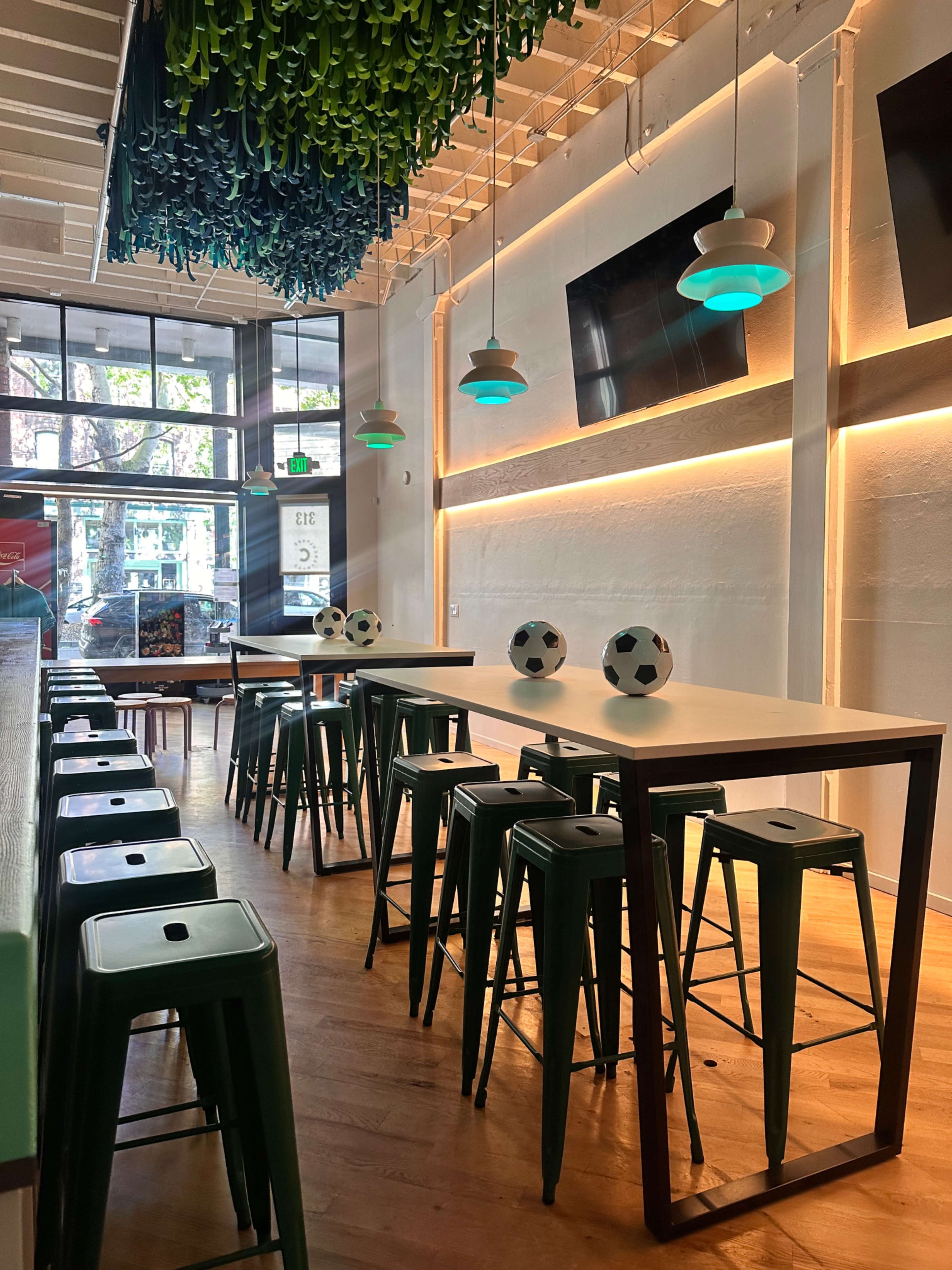 The image shows a modern café or bar with a long table surrounded by high stools, soccer-themed decor, and pendant lights hanging from the ceiling.