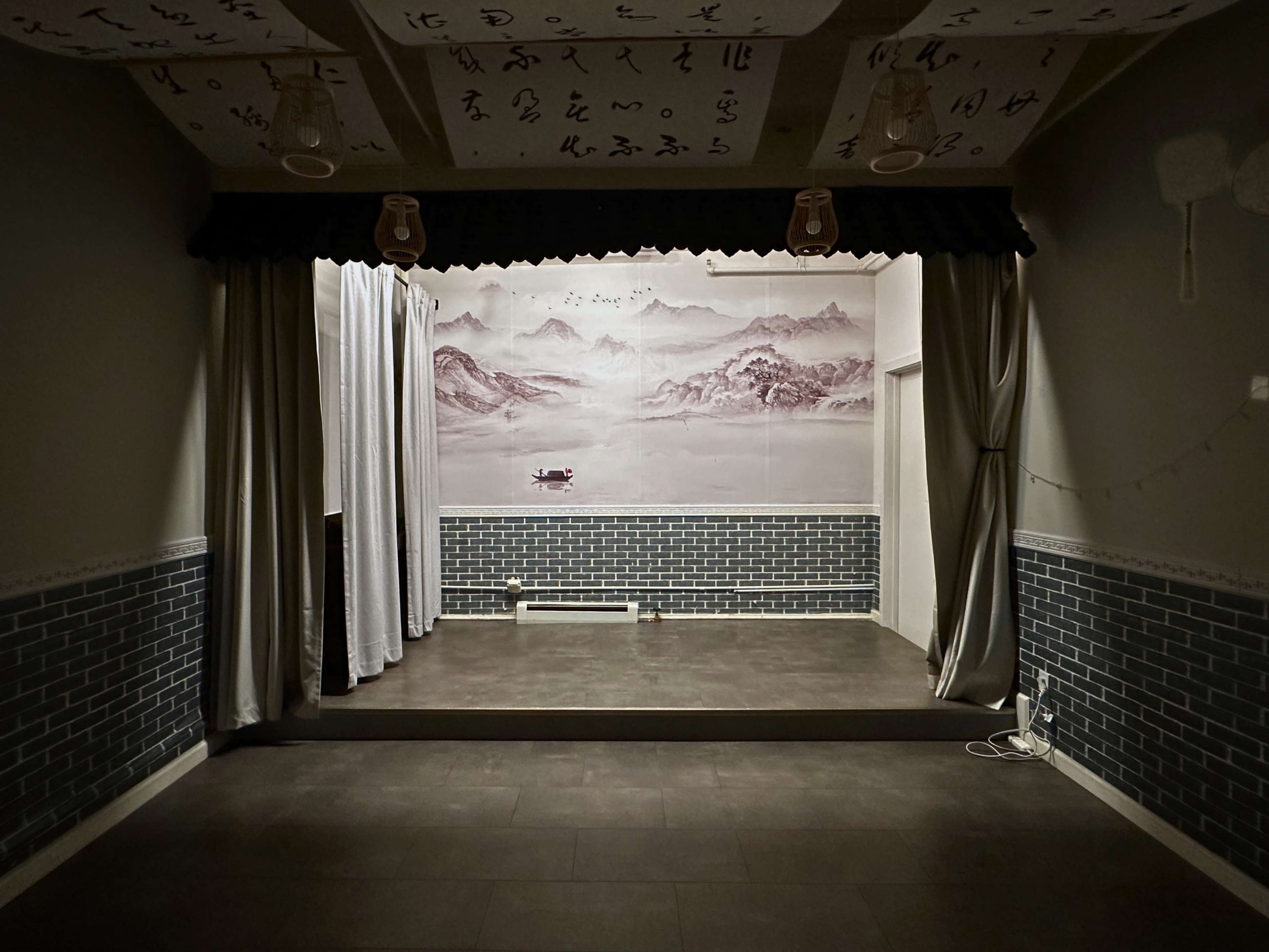 The image shows an empty stage area with a mural of mountains and a boat in the background, framed by white curtains and a tiled wall.