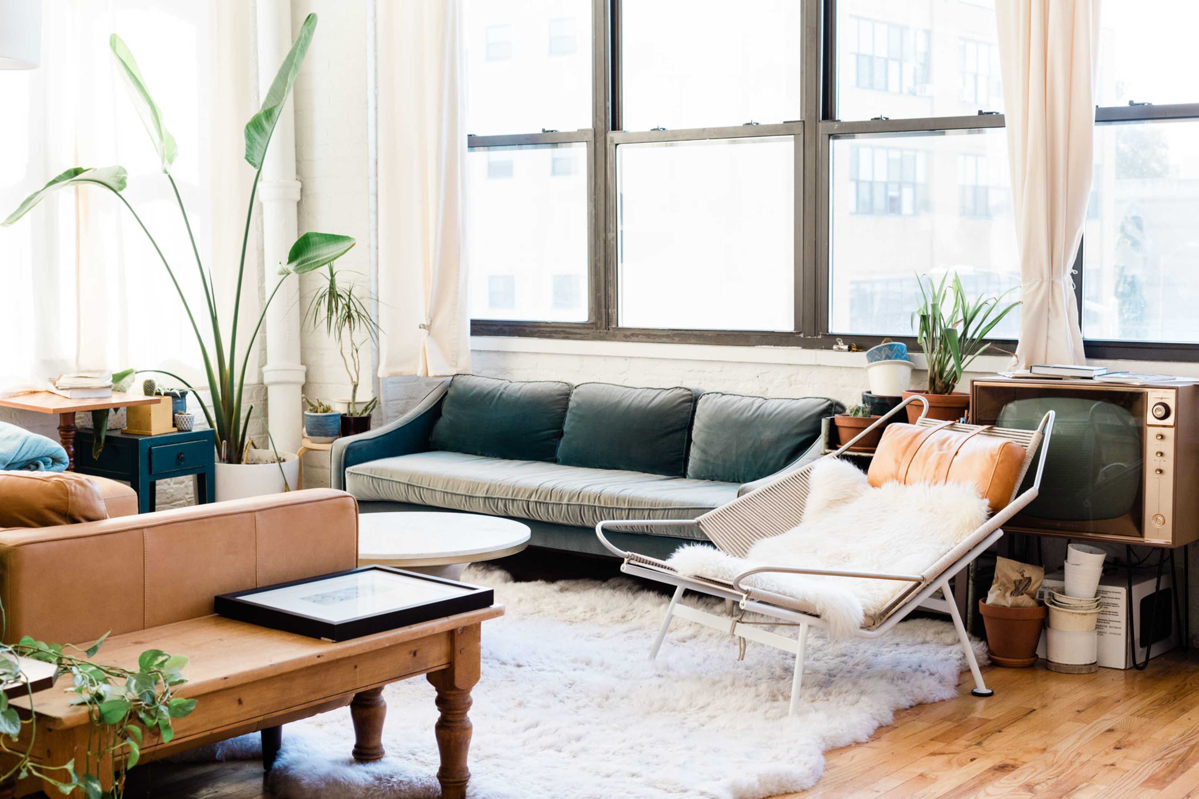 The image depicts a bright living room featuring a green sofa, a white lounge chair with a fur throw, a coffee table, and large windows framing a view of a cityscape.