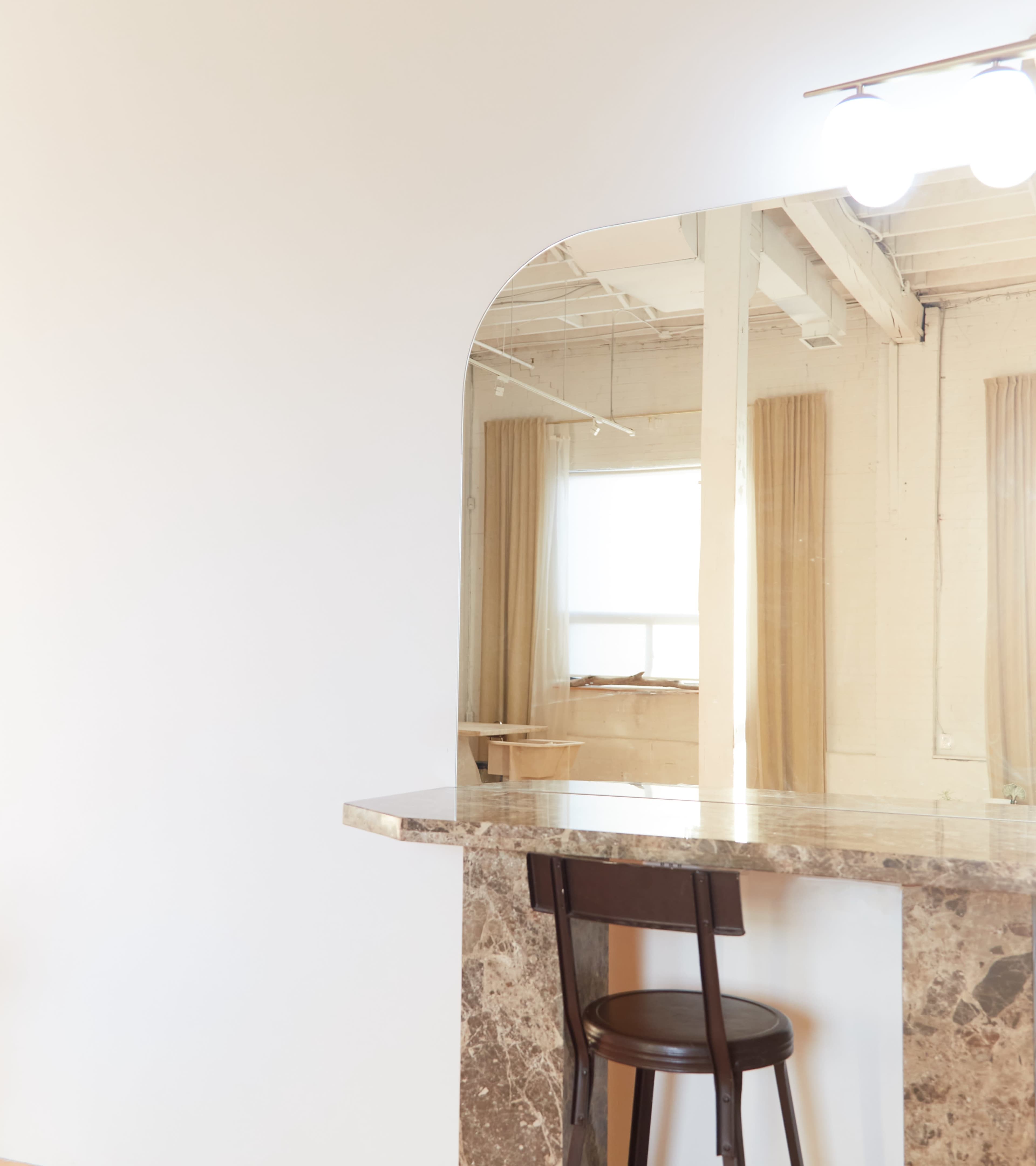A minimalist interior features a marble countertop with a single wooden chair and a large arched mirror above it.