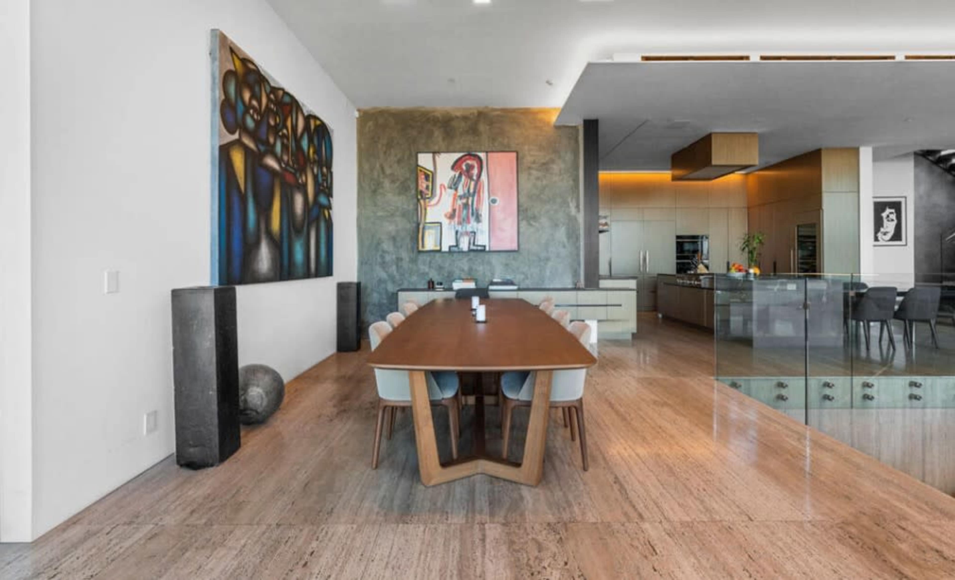 A modern dining area features a long wooden table surrounded by upholstered chairs, with large artworks on the walls and a sleek kitchen visible in the background.