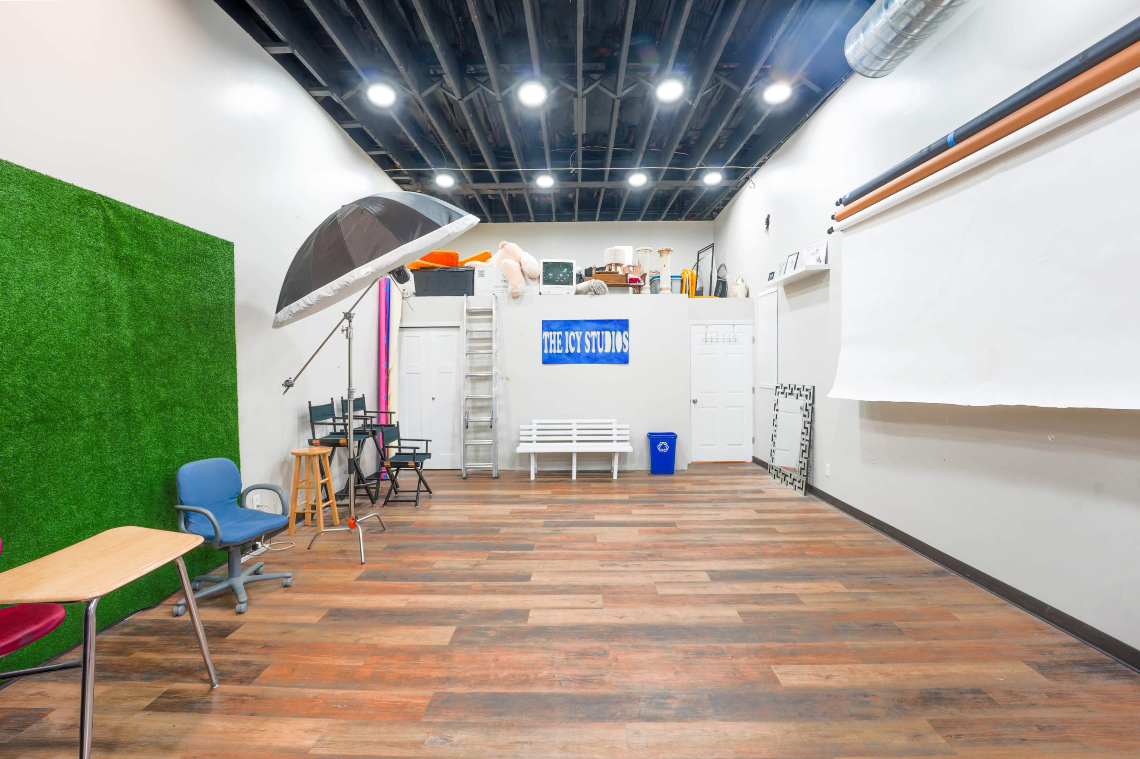 The image depicts a photography studio featuring wooden flooring, a large umbrella light, various props, and a backdrop area with green grass-like material.