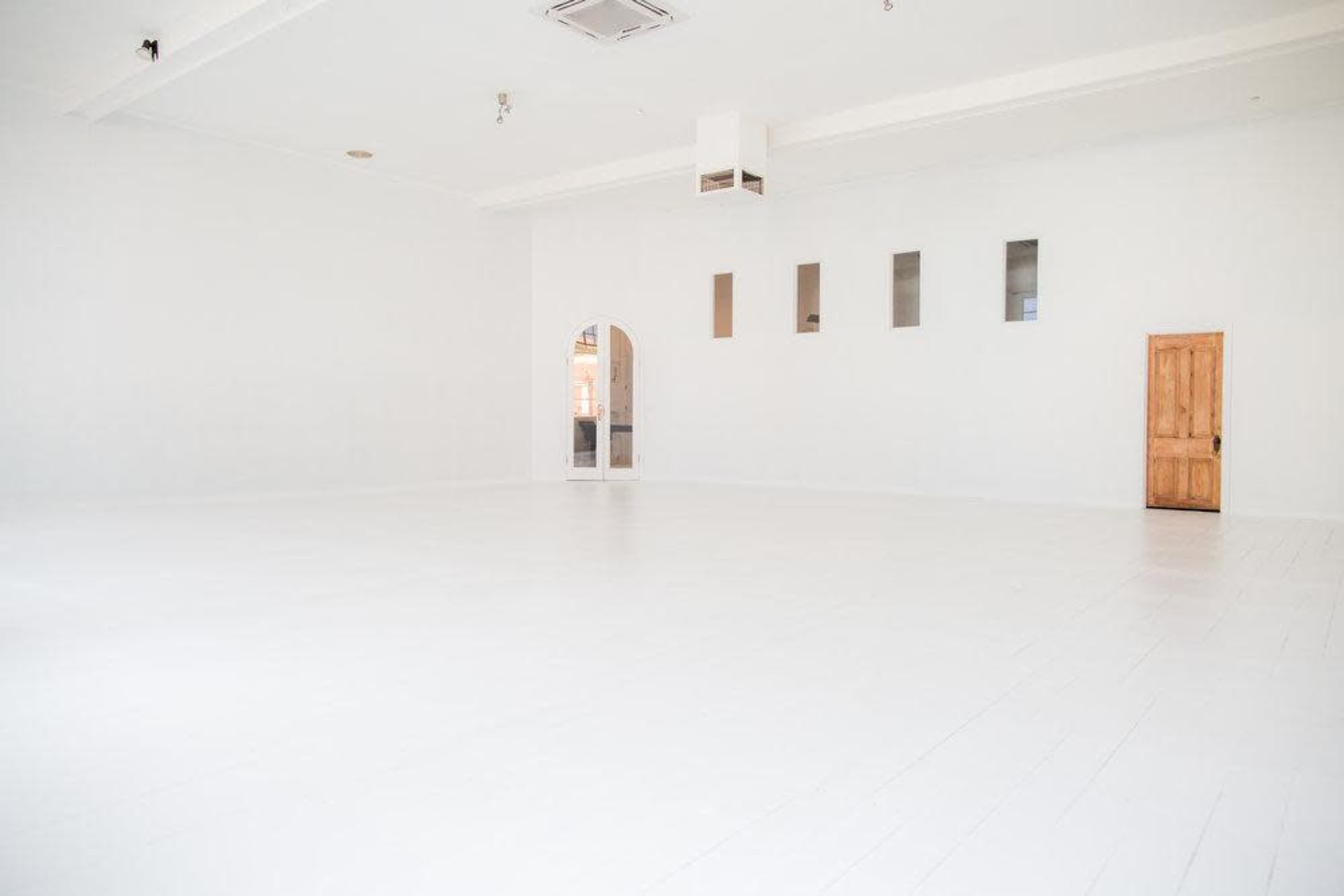 The image shows a spacious, empty room with white walls, a wooden door, and several small windows.