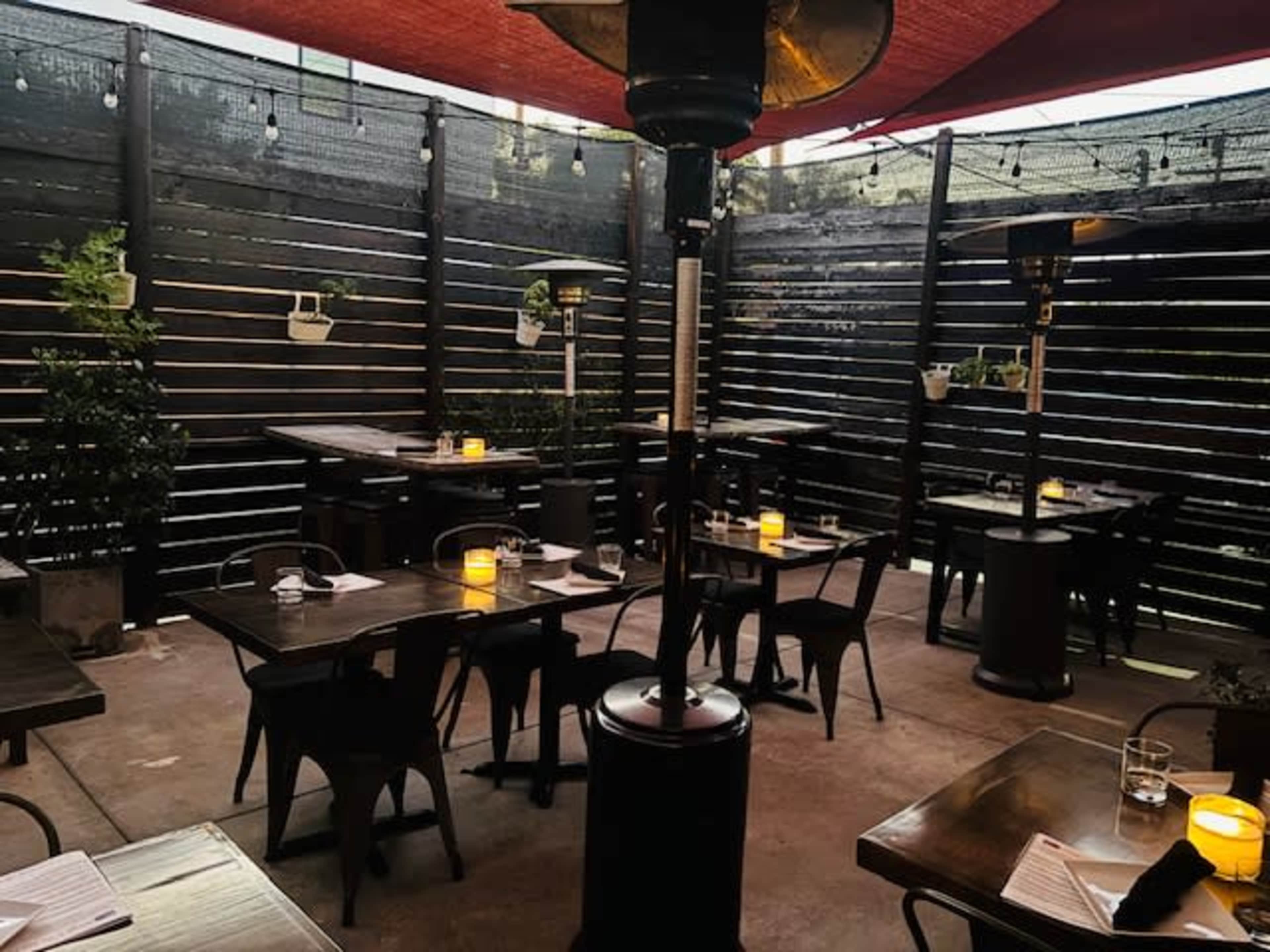 The image shows a cozy outdoor restaurant patio with wooden tables, metal chairs, and warm lighting from candlelit centerpieces under a shaded canopy.