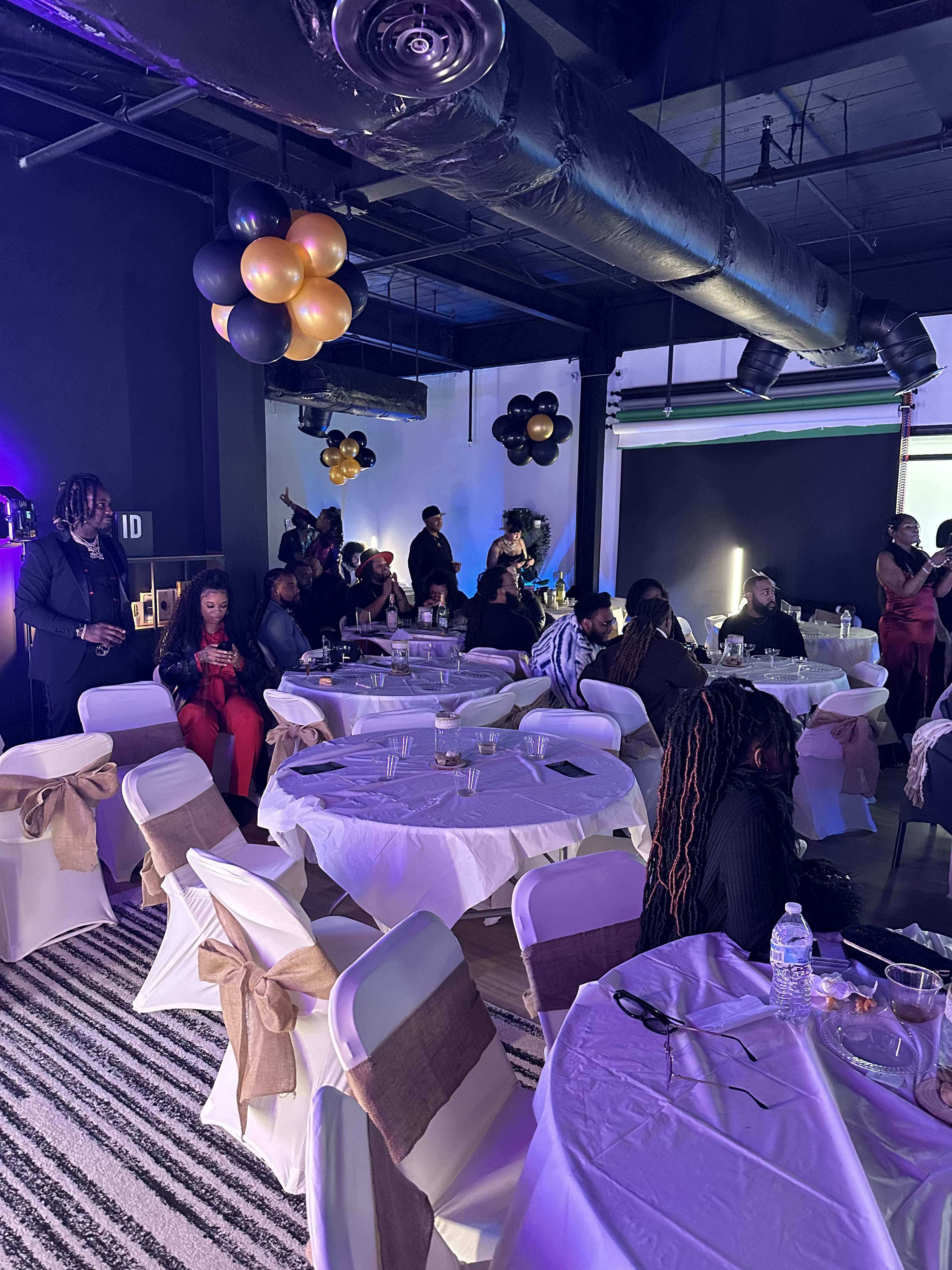 A gathering is taking place in a decorated venue with tables covered in white tablecloths and adorned with balloons in gold and black.