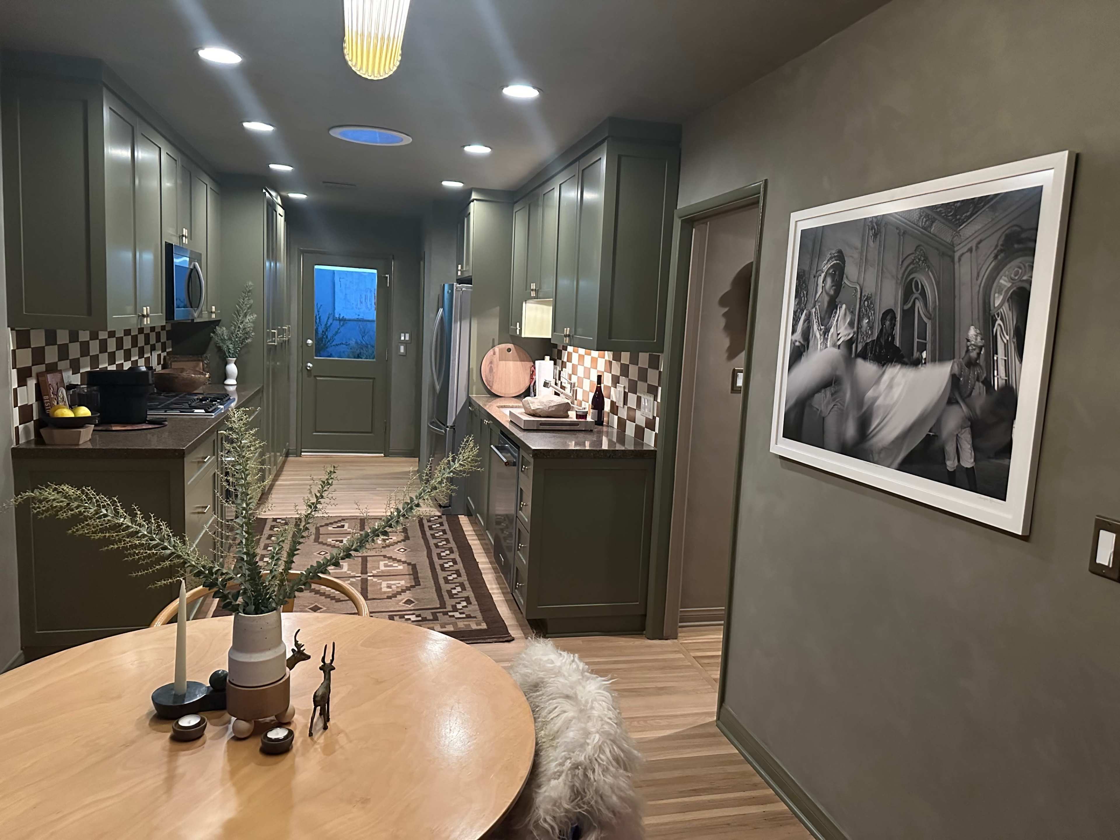 The image shows a modern kitchen with green cabinetry, a wooden dining table, and a large framed black-and-white photograph on the wall.