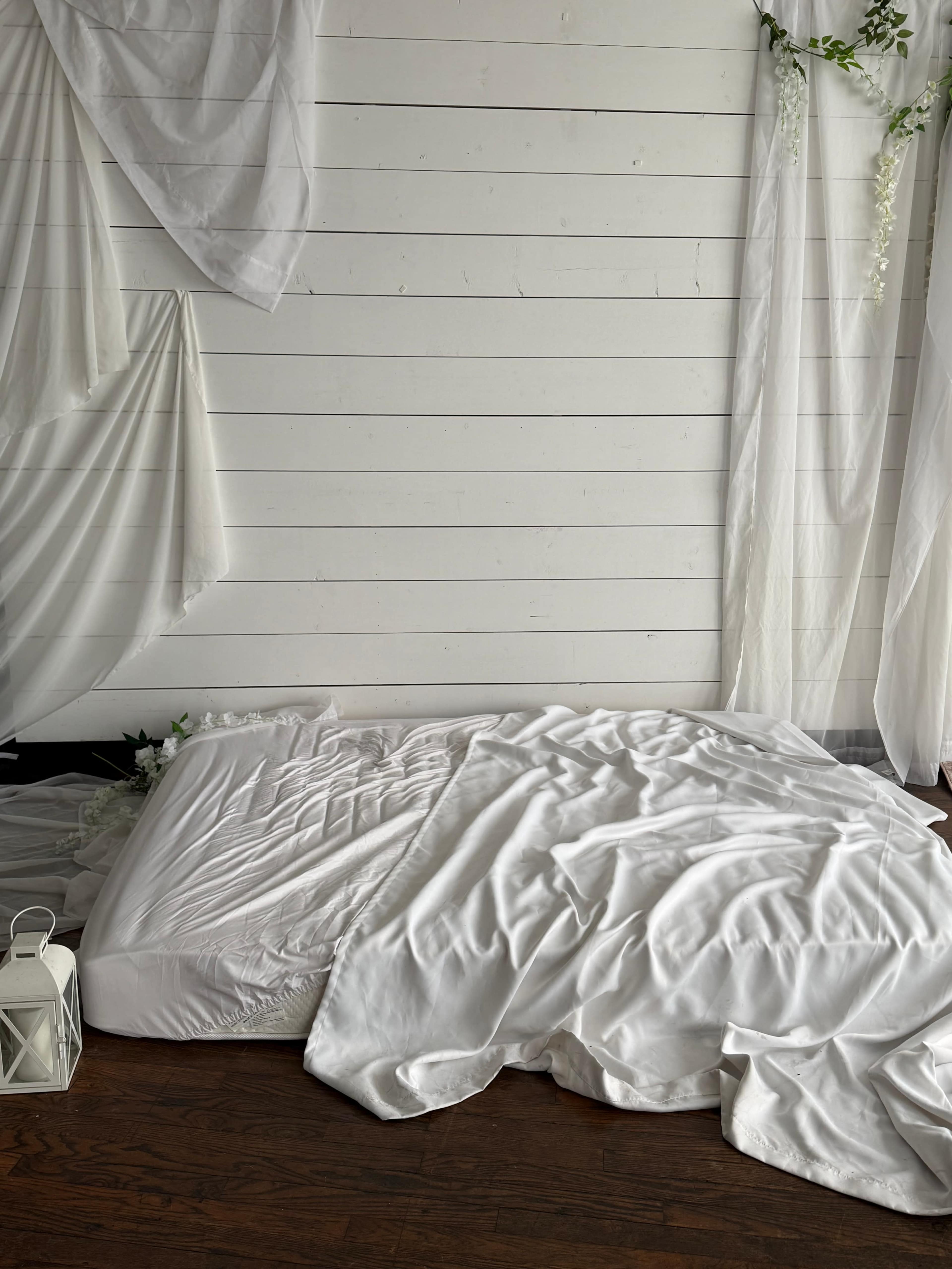 A neatly unmade bed with a crumpled white duvet on a wooden floor, surrounded by white curtains and floral decorations.