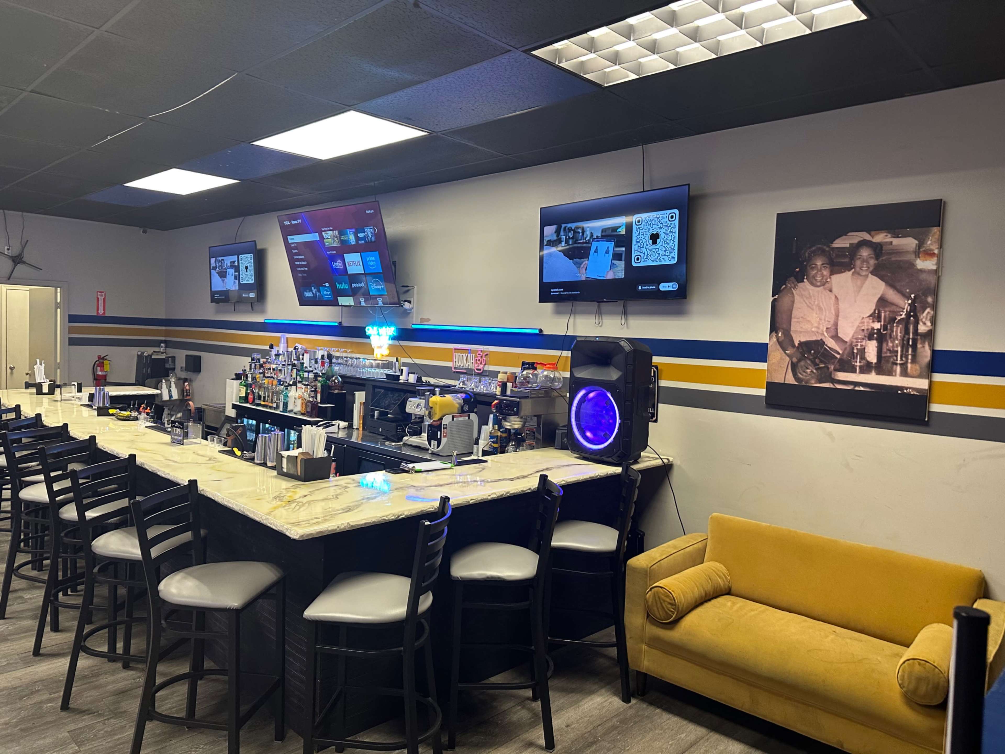 The image shows a bar area with a curved counter, high stools, two large screens on the wall, and a yellow couch beside a framed black-and-white photo.