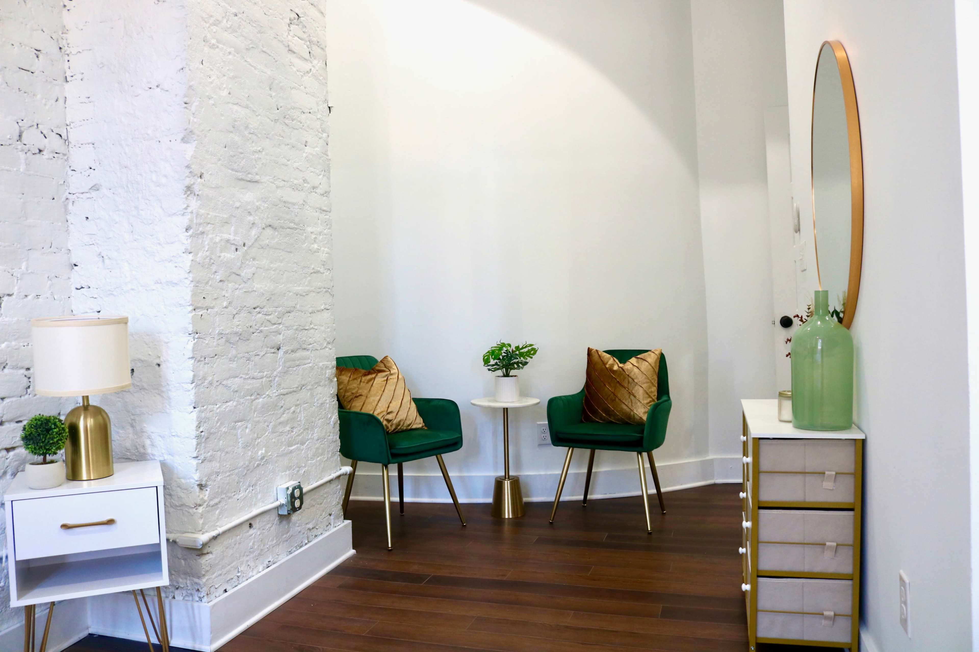 A corner of a modern room features two green chairs with gold accents, a small table between them, and a decorative mirror on the wall.