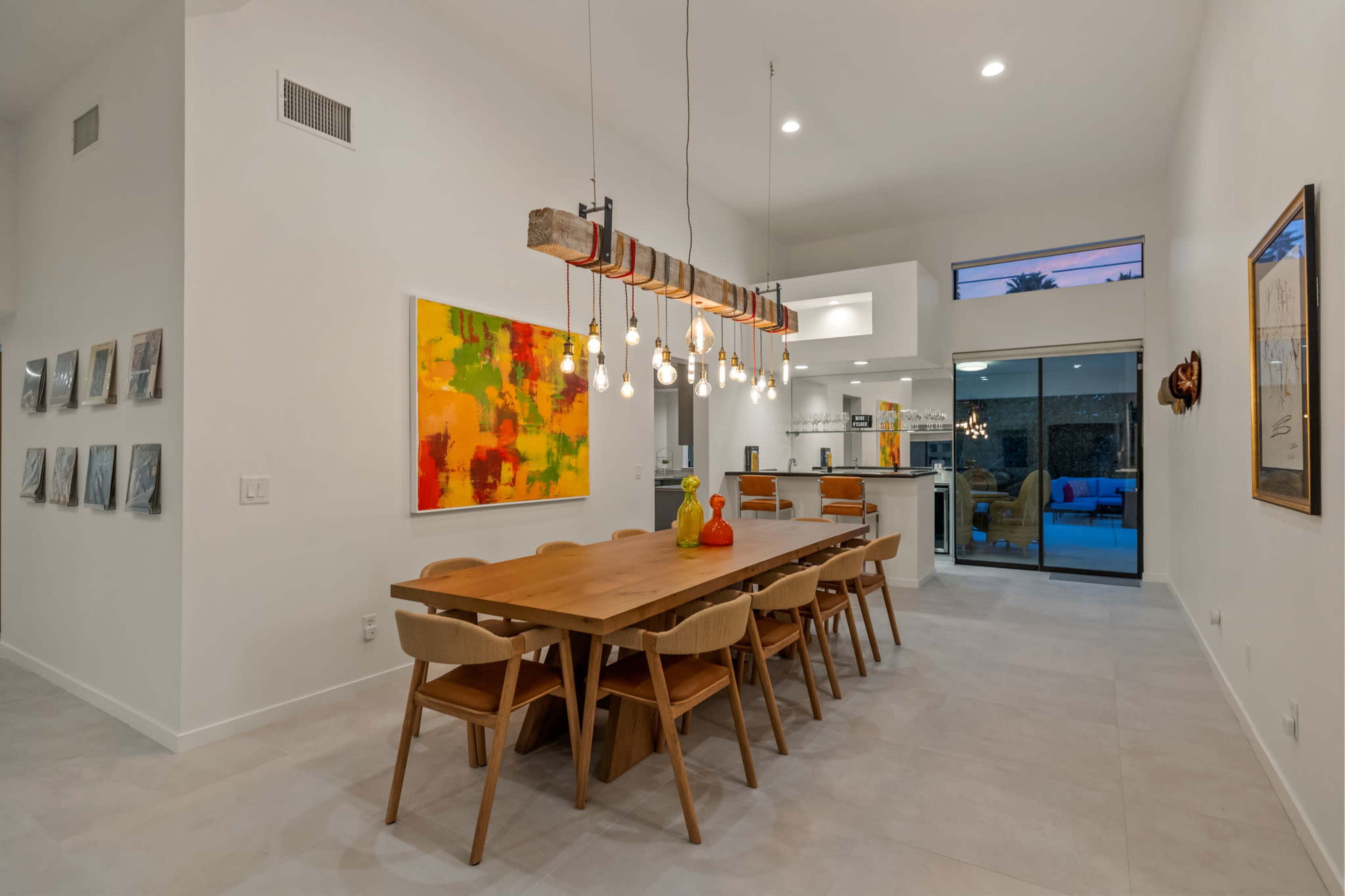 The image shows a modern dining area with a large wooden table, surrounded by chairs, and a brightly colored abstract painting on the wall.