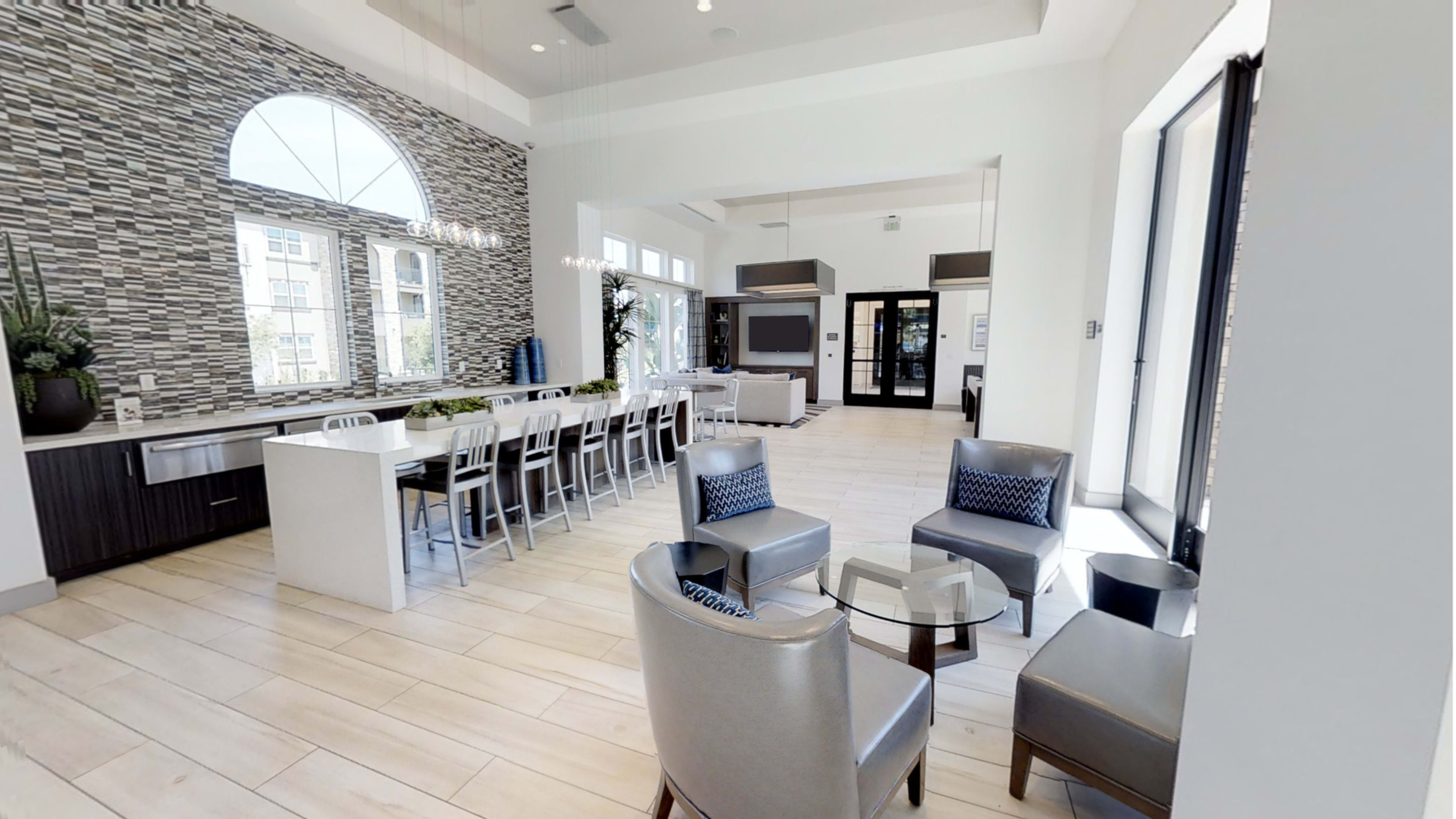 A modern communal space with a long table and chairs, a lounge area featuring grey armchairs, and large windows letting in natural light.