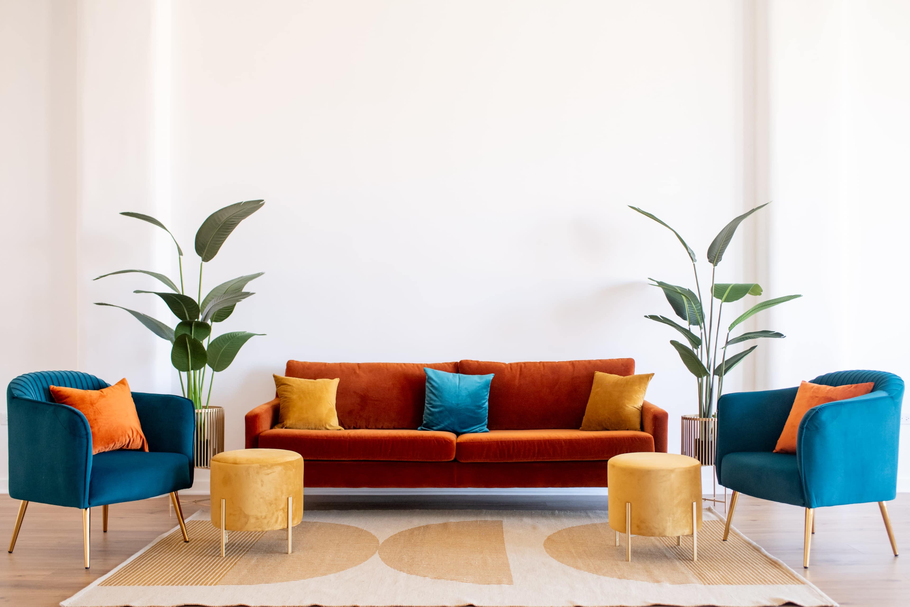 A living room with an orange couch, two turquoise armchairs, and round stools, accompanied by potted plants against a white wall.