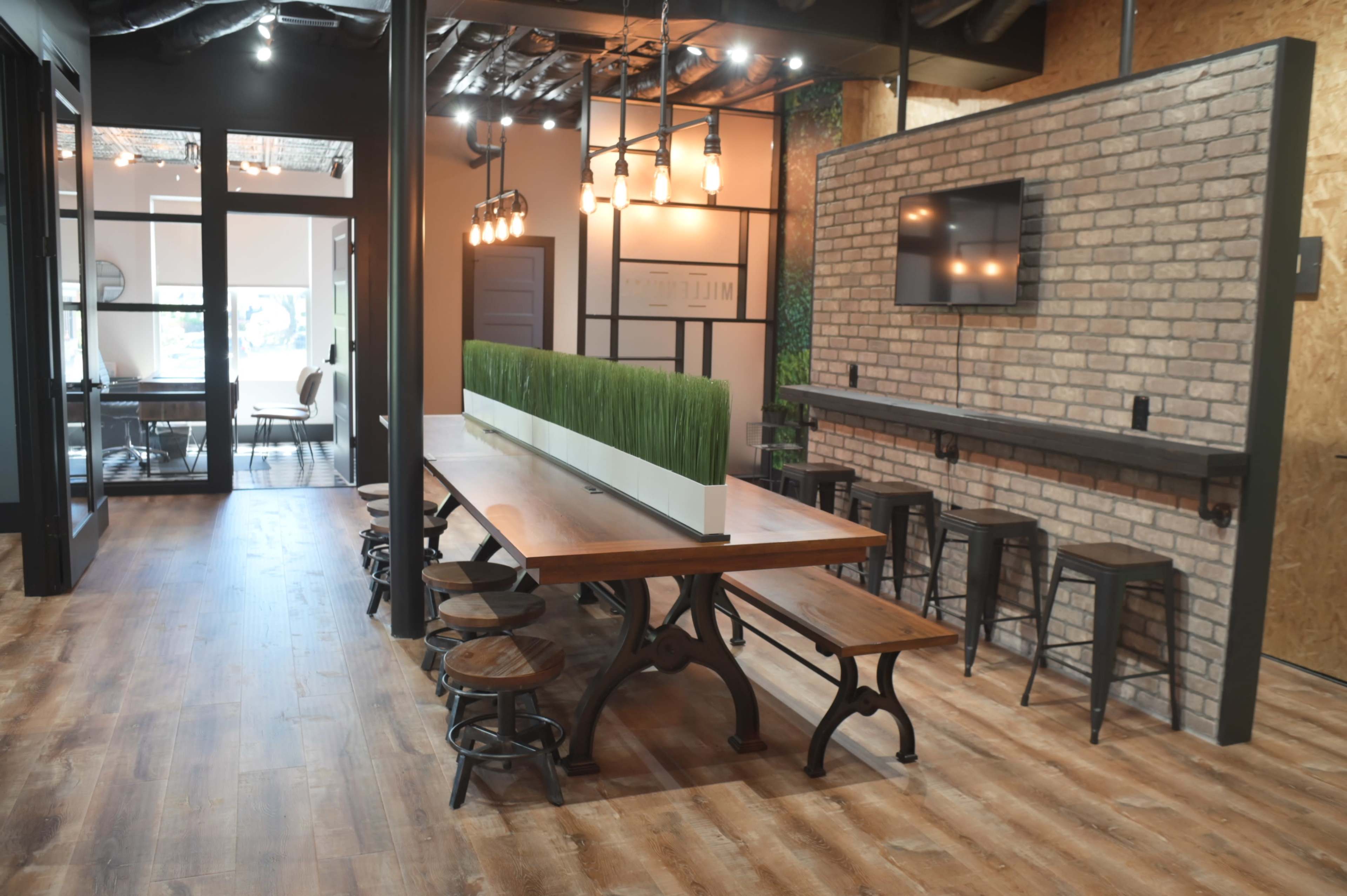 A modern office interior featuring a long wooden table with benches, surrounded by industrial-style lighting and a brick accent wall with a television.