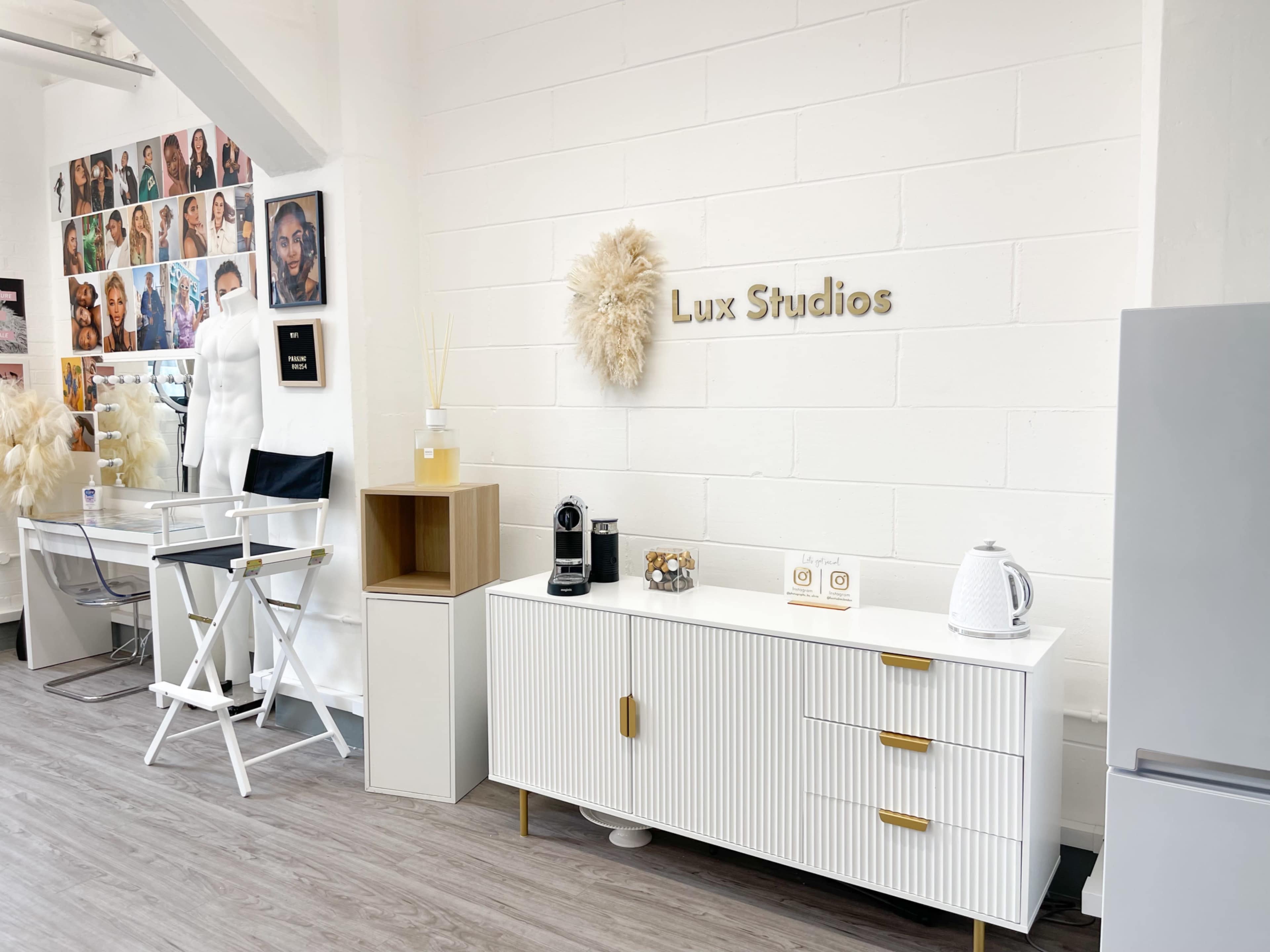 A modern studio interior featuring a white wall with a decorative sign reading "Lux Studios," along with a cabinet, a coffee machine, and a gallery of portraits on the adjacent wall.