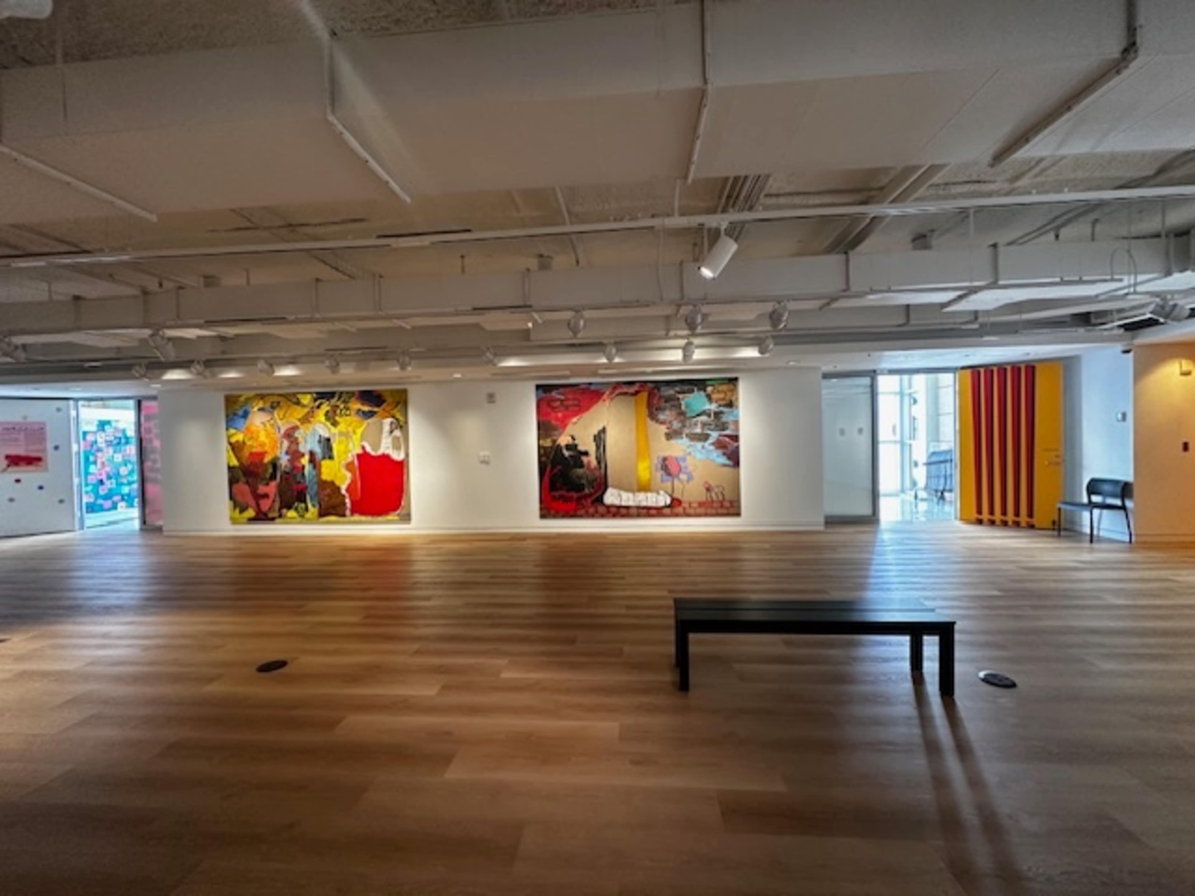 An art gallery with two large murals on one wall and a wooden bench in the center of the room, featuring a light wood floor and modern lighting.