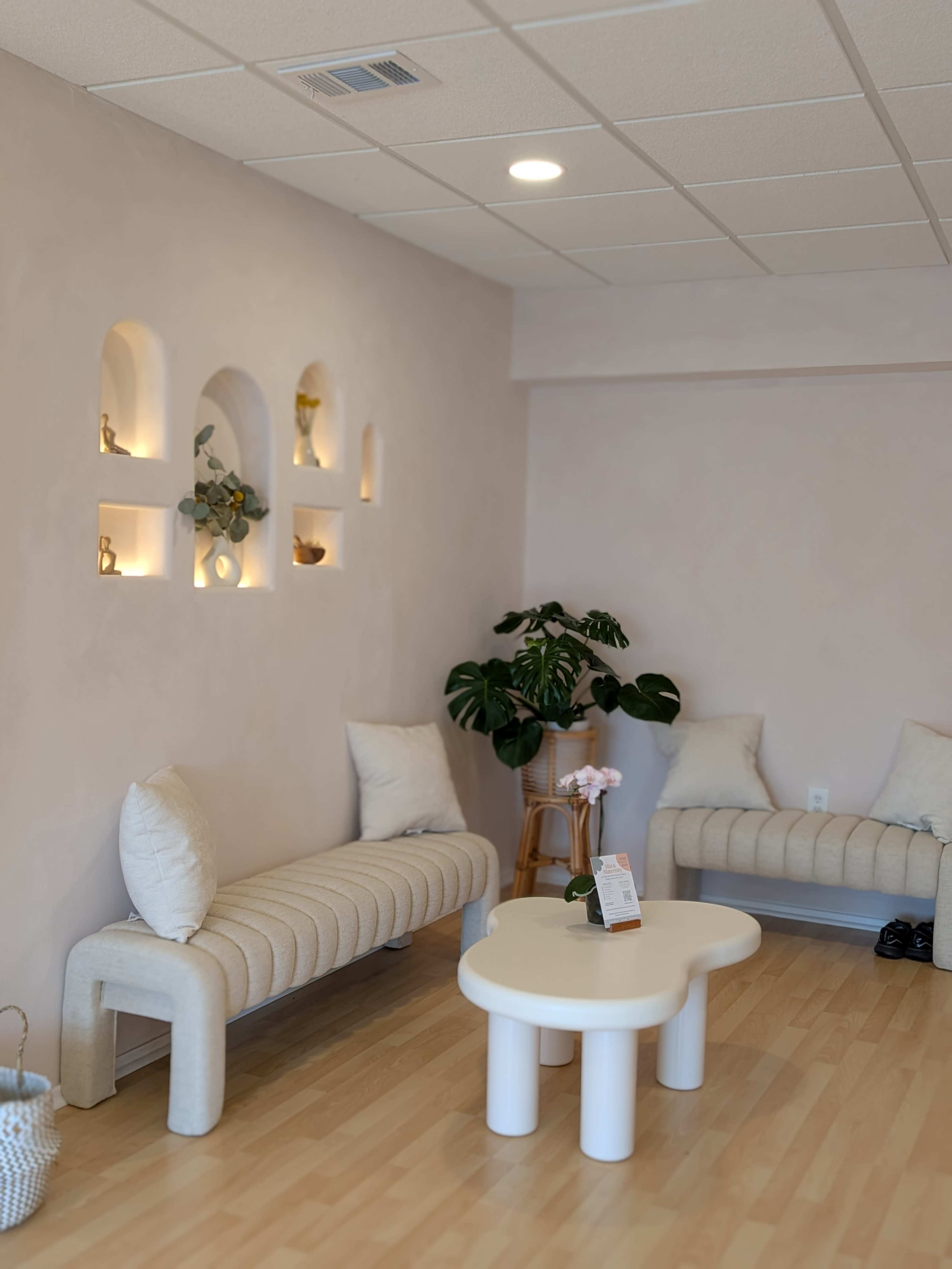 The image shows a softly lit interior of a cozy waiting area featuring two curved benches, a round table, and decorative niches on the wall.