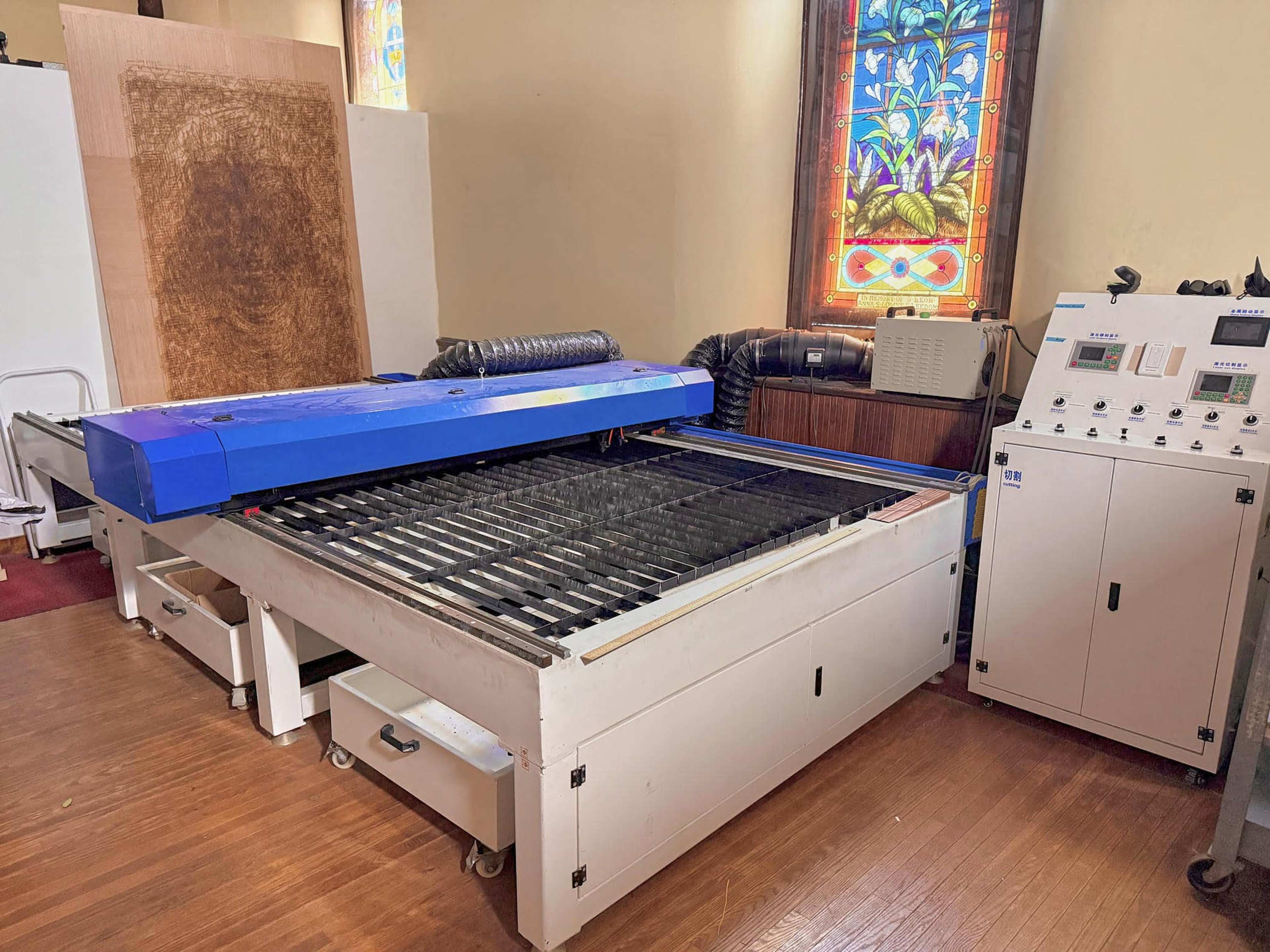 The image shows a large laser cutting machine with a blue cover, situated in a room featuring a stained glass window.