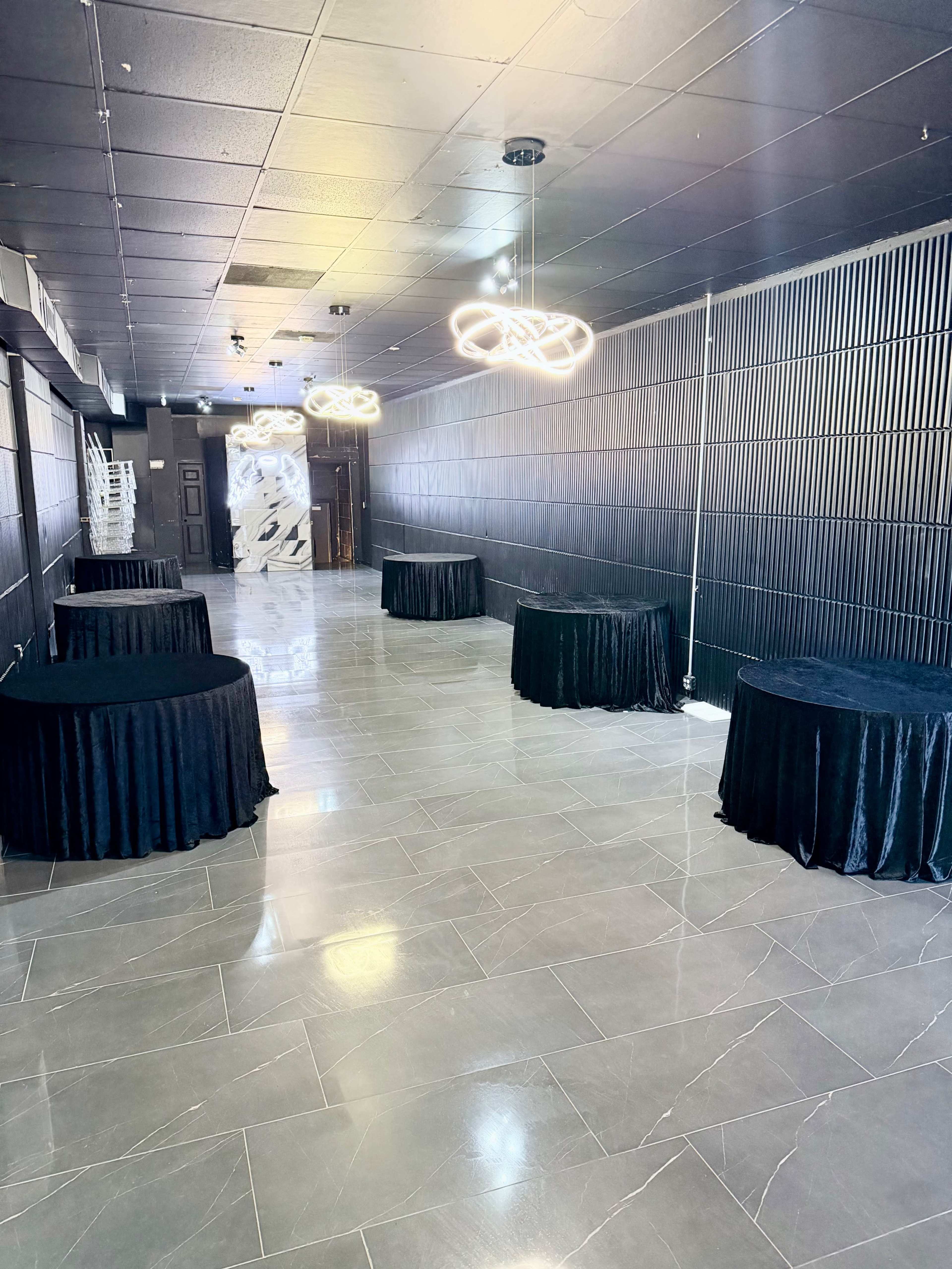 The image shows a spacious room with several round tables covered in black cloth and modern light fixtures hanging from the ceiling.